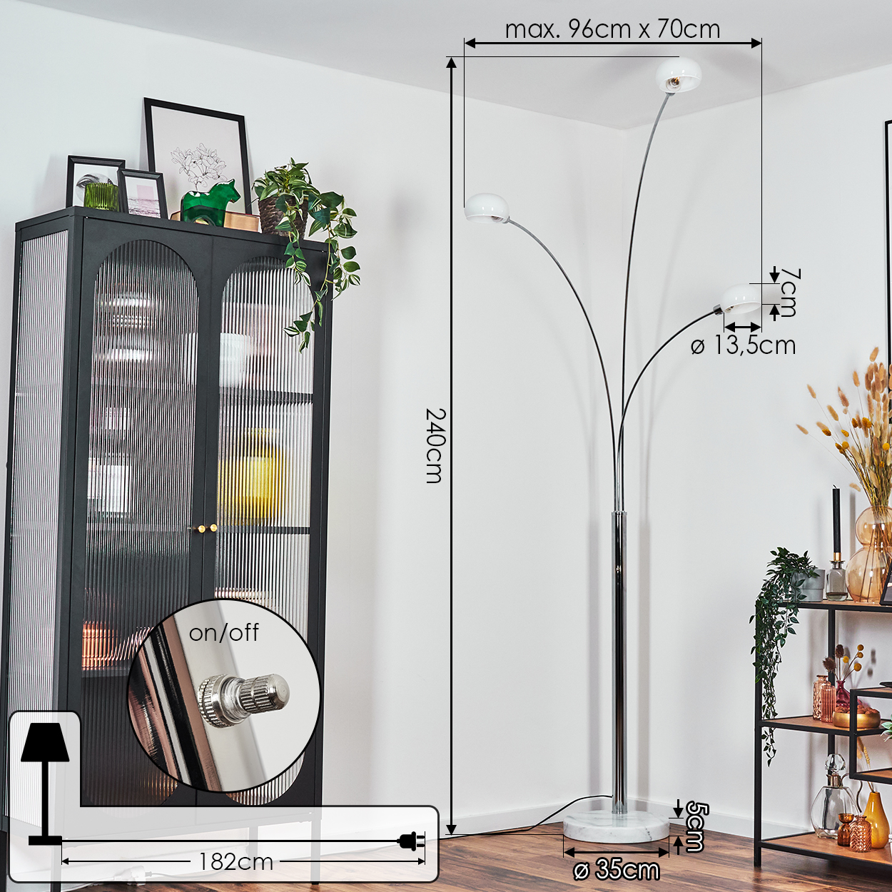 Eine moderne Stehlampe mit drei Lichtern steht neben einem schwarzen Schrank mit Pflanzen und Dekorationen. Die Wand ist weiß.