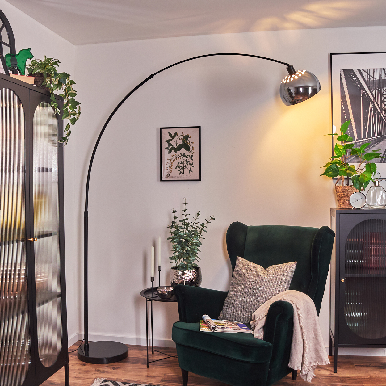 Ein dunkelgrüner Sessel mit Decke und Kissen. Eine schwarze Bogenlampe und ein schwarzer Schrank mit Pflanzen und Bildern sind sichtbar.