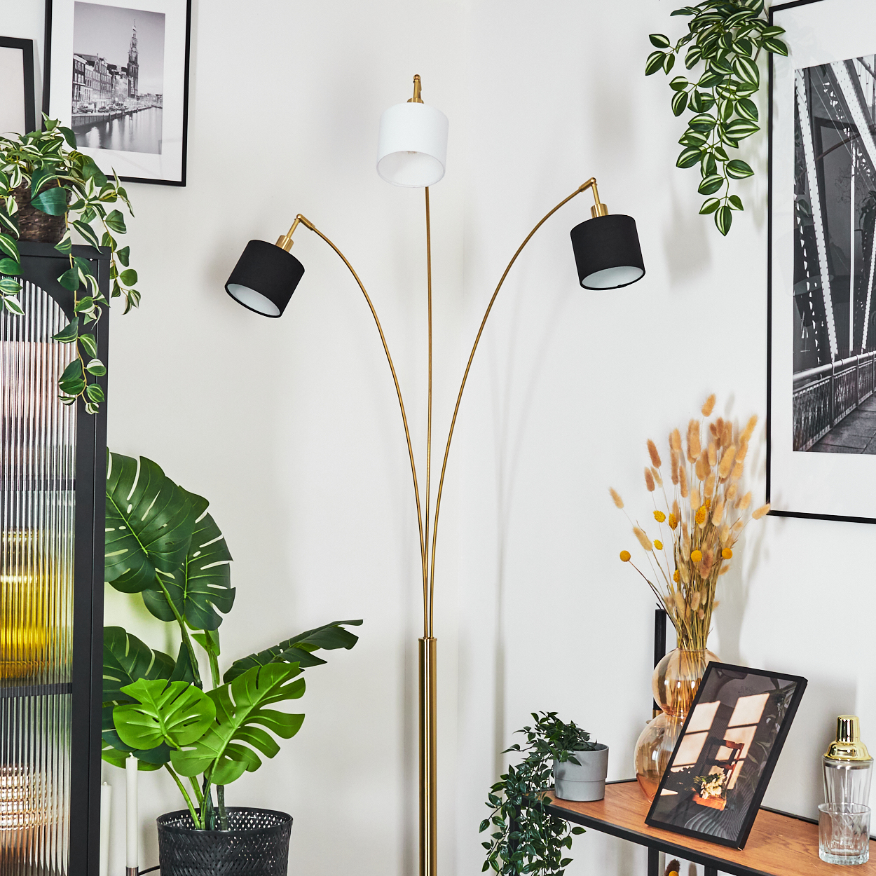 Goldene Stehlampe mit drei Köpfen. Pflanzen und Bilder schmücken die weißen Wände. Ein schwarzes Regal hält Deko.
