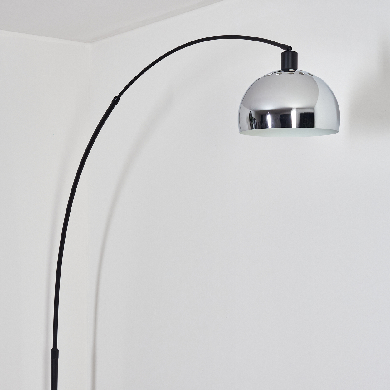 Eine moderne Bogenlampe mit verchromtem Schirm und schwarzem Rahmen steht vor einer weißen Wand. Die Lampe ist aus.