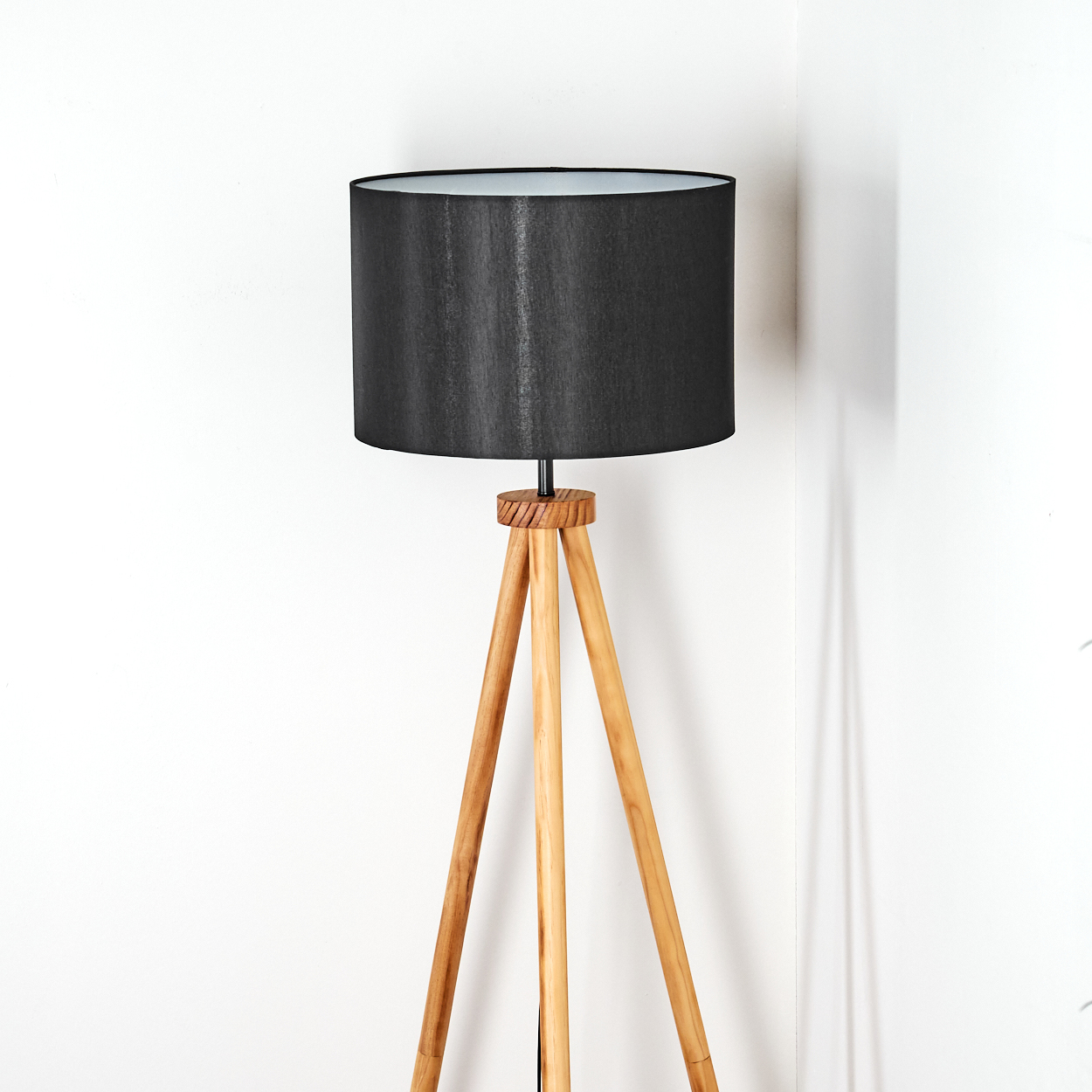 Eine Stehlampe mit schwarzem Lampenschirm und Holzstativbeinen steht vor einer weißen Wand. Die Lampe steht in der Ecke eines Raumes.