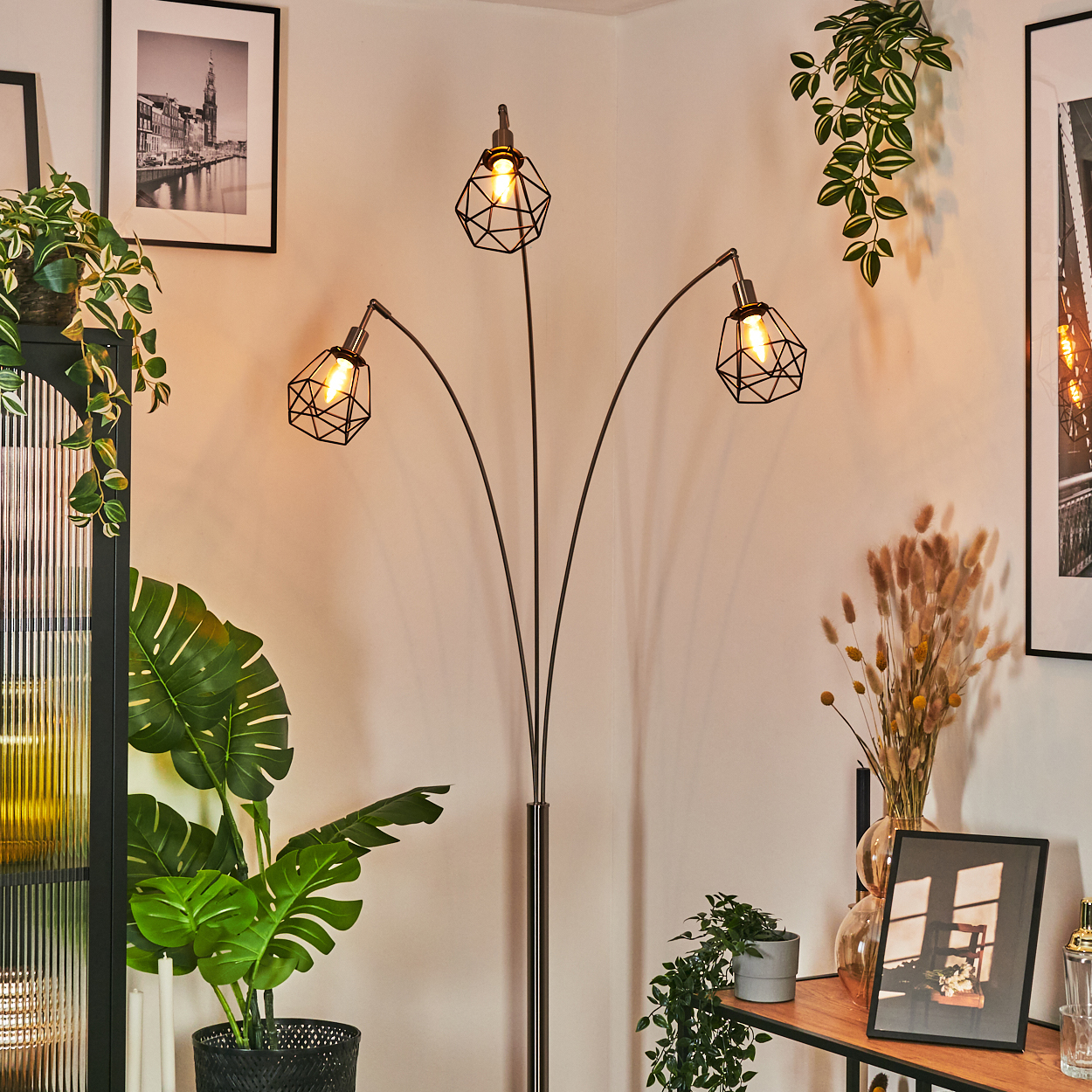 Eine moderne Stehlampe mit drei käfigförmigen Leuchten. Pflanzen und Bilder schmücken den Raum.