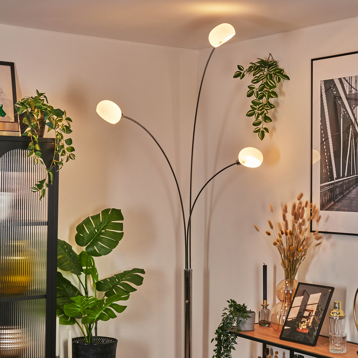 Ein modernes Wohnzimmer mit Stehlampe, Pflanzen und Dekoration. Die Lampe hat vier Lichter. An der Wand befindet sich auch ein Bild.