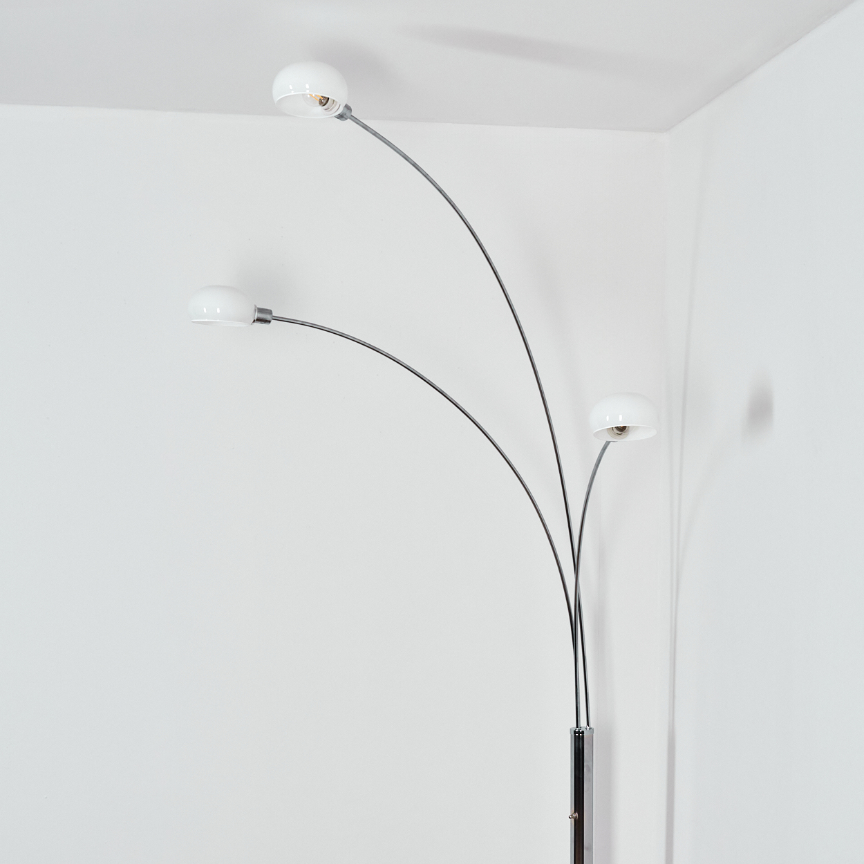 Eine moderne Stehlampe mit drei weißen Lampenschirmen auf gebogenen Metallarmen vor einer weißen Wand.