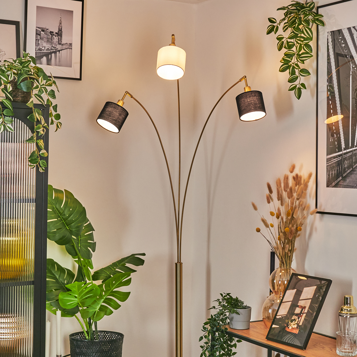 Eine moderne Stehlampe mit drei Lichtern steht in einer Zimmerecke. Pflanzen, gerahmte Bilder und ein Regal mit Dekorationen sind vorhanden.