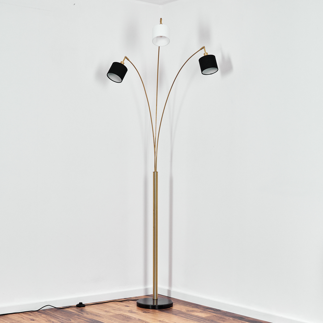 Eine moderne Stehlampe mit drei Köpfen. Zwei schwarze und ein weißer Schirm. Gold- und Schwarzbasis. Steht in einer Raumecke.