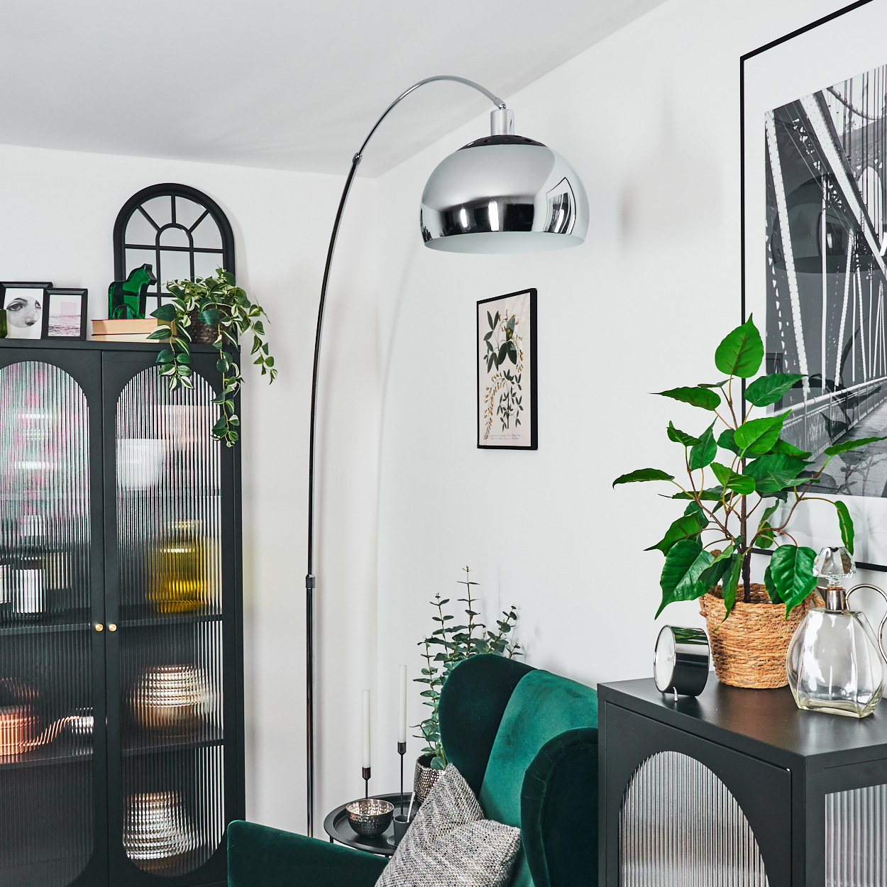 Ein modernes Wohnzimmer mit einer großen Bogenlampe, grünen Pflanzen, einem schwarzen Schrank und einem grünen Samtarmstuhl. Ein Brückenfoto hängt an der Wand.