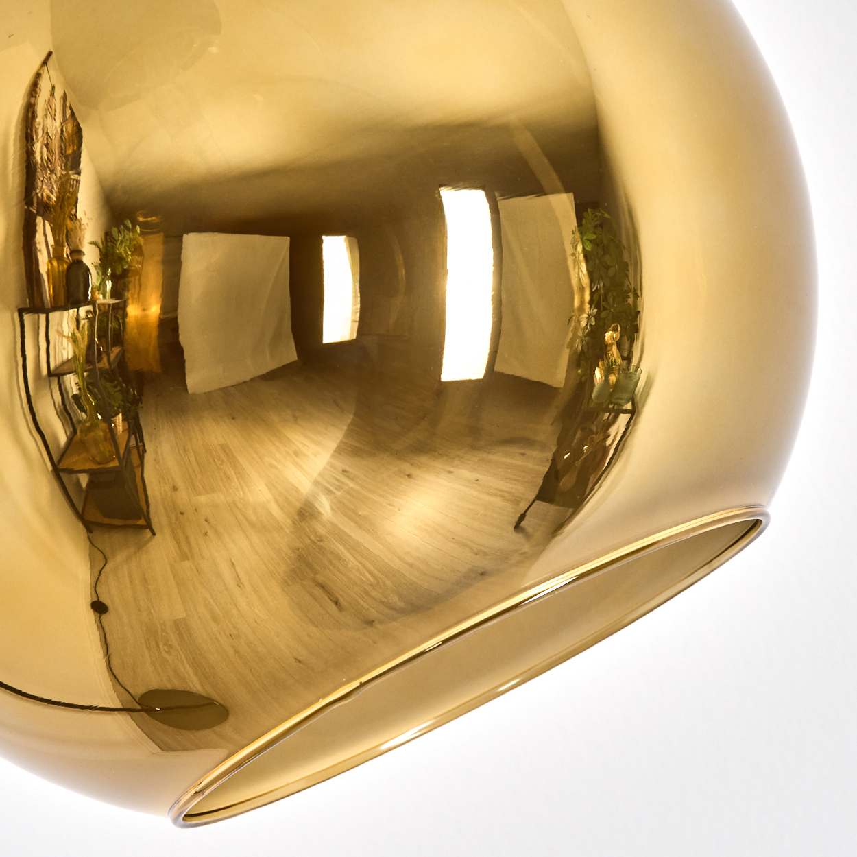 Eine goldene, kugelförmige Lampe spiegelt einen Raum mit Fenstern, Pflanzen und Holzböden wider. Die Lampe steht vor weißem Hintergrund.
