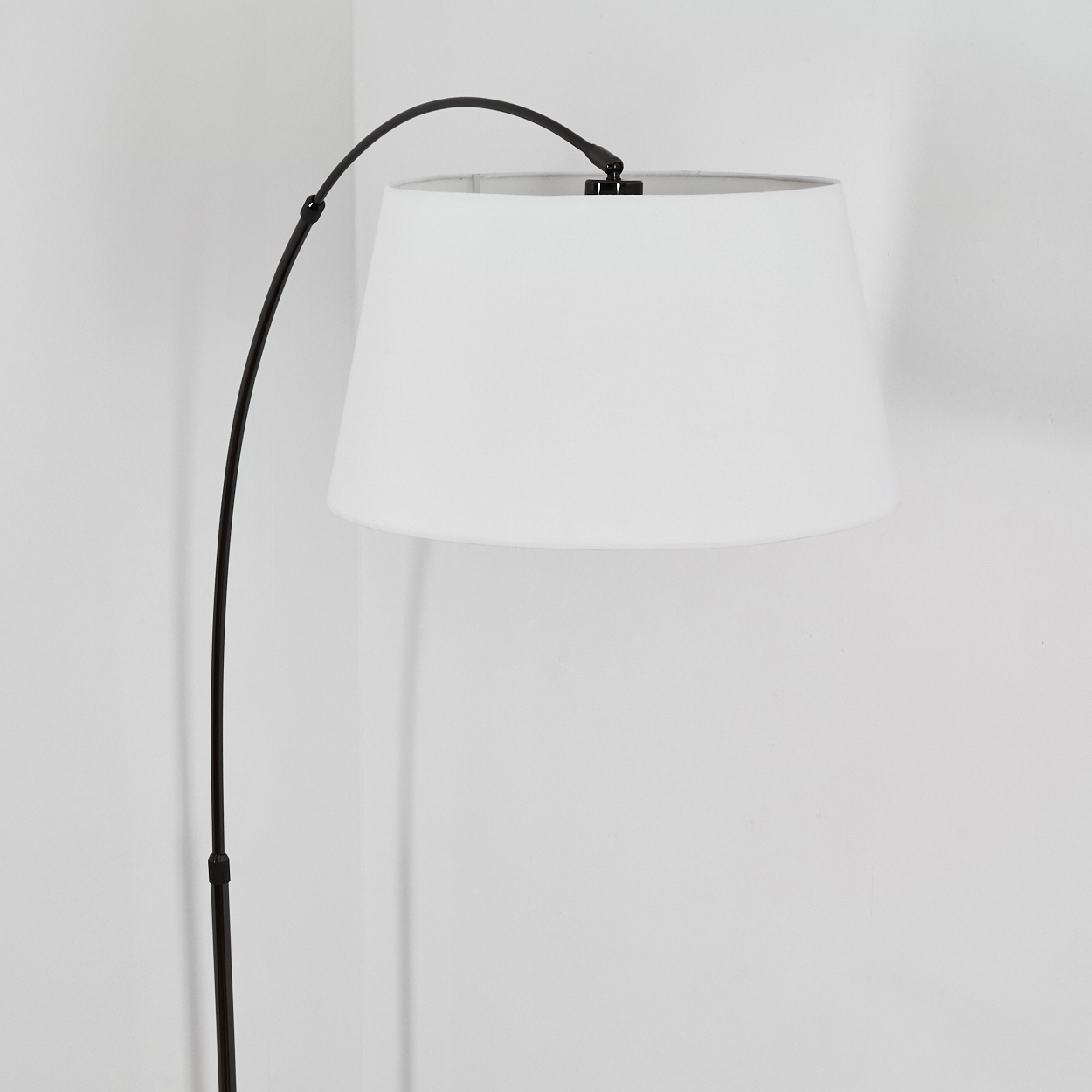 Eine moderne Stehlampe. Sie hat einen schwarzen, gebogenen Arm, der einen weißen Lampenschirm hält. Der Hintergrund ist eine weiße Wand.
