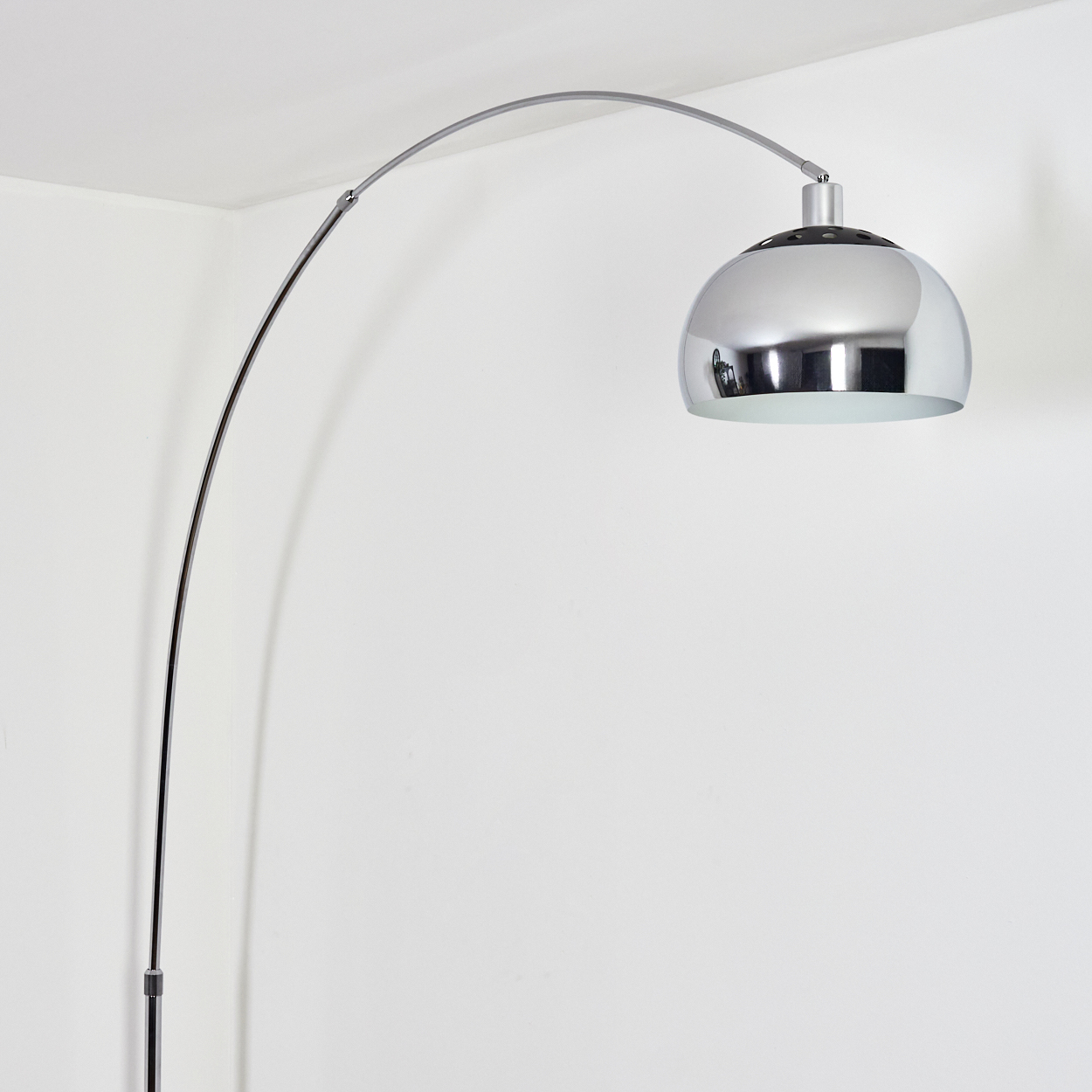 Eine verchromte Bogenlampe mit gewölbtem Schirm. Sie steht in einer weißen Ecke und spiegelt den Raum wider. Die Lampe ist ein modernes Design.