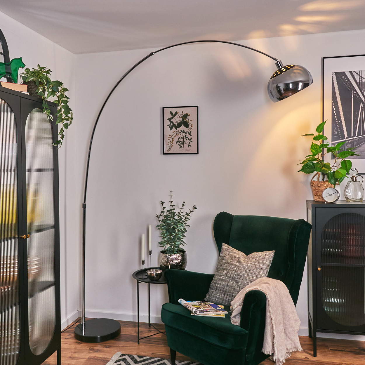 Ein modernes Wohnzimmer mit grünem Sessel, Stehlampe und schwarzem Schrank. Pflanzen und Deko sorgen für eine gemütliche Atmosphäre.