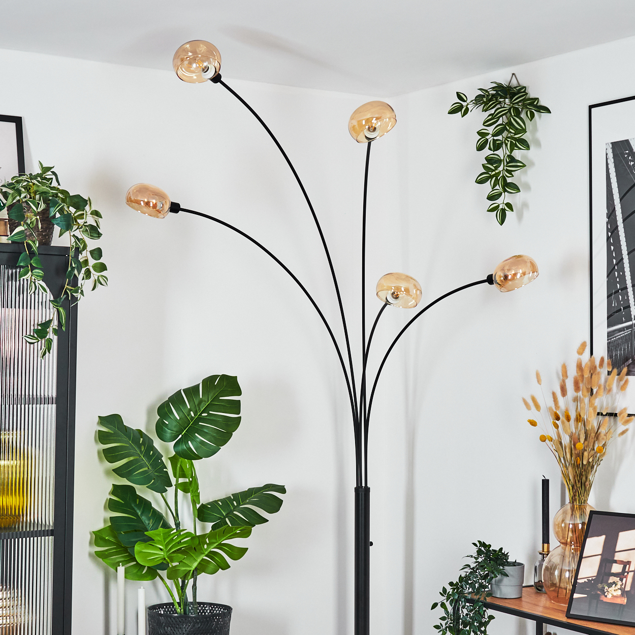 Eine moderne Stehlampe mit fünf bernsteinfarbenen Glasschirmen, schwarzem Rahmen, steht neben Topfpflanzen und Wanddekoration.