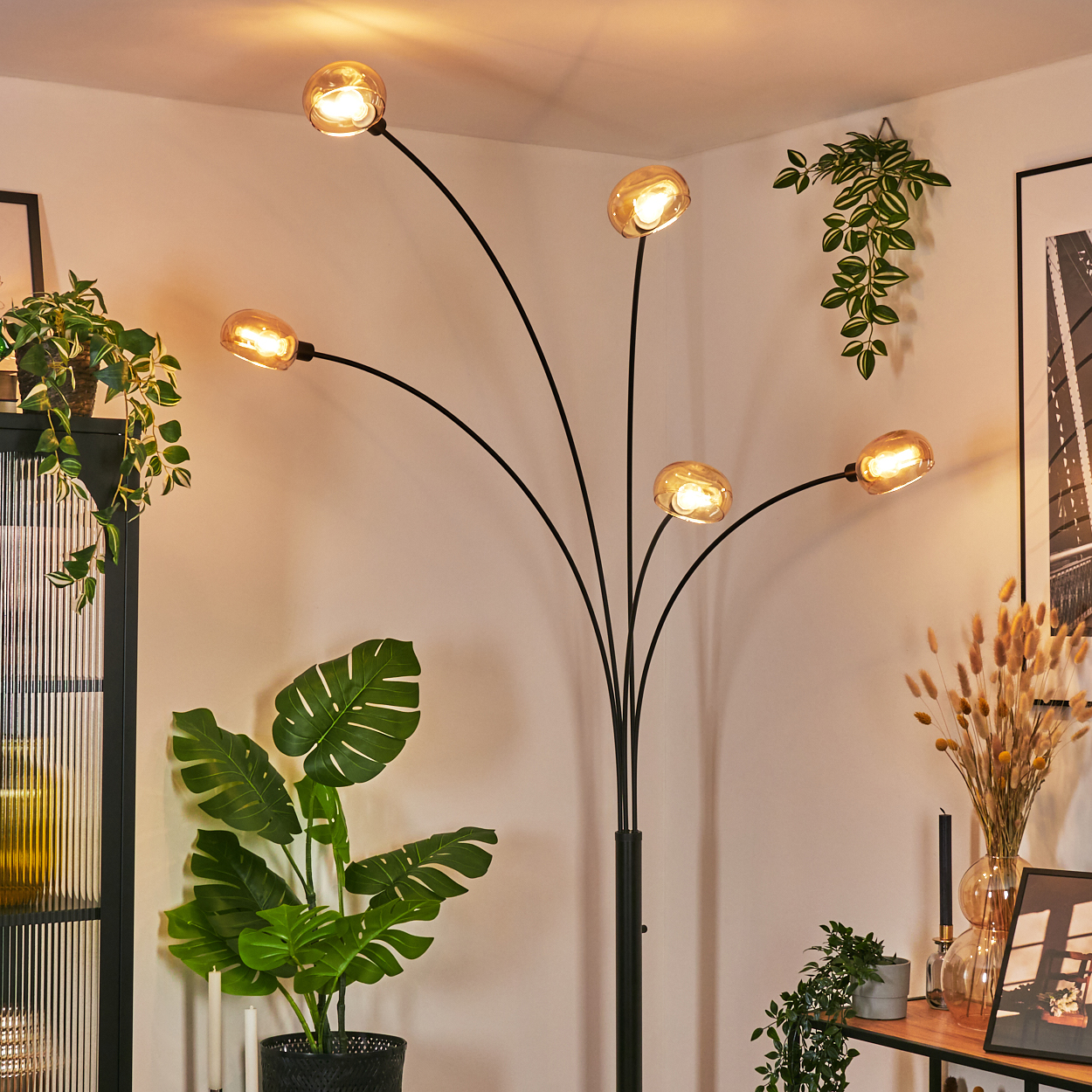 Eine moderne Stehlampe mit fünf Lichtern. Pflanzen und Dekorationen umgeben sie in einem Raum mit hellen Wänden.