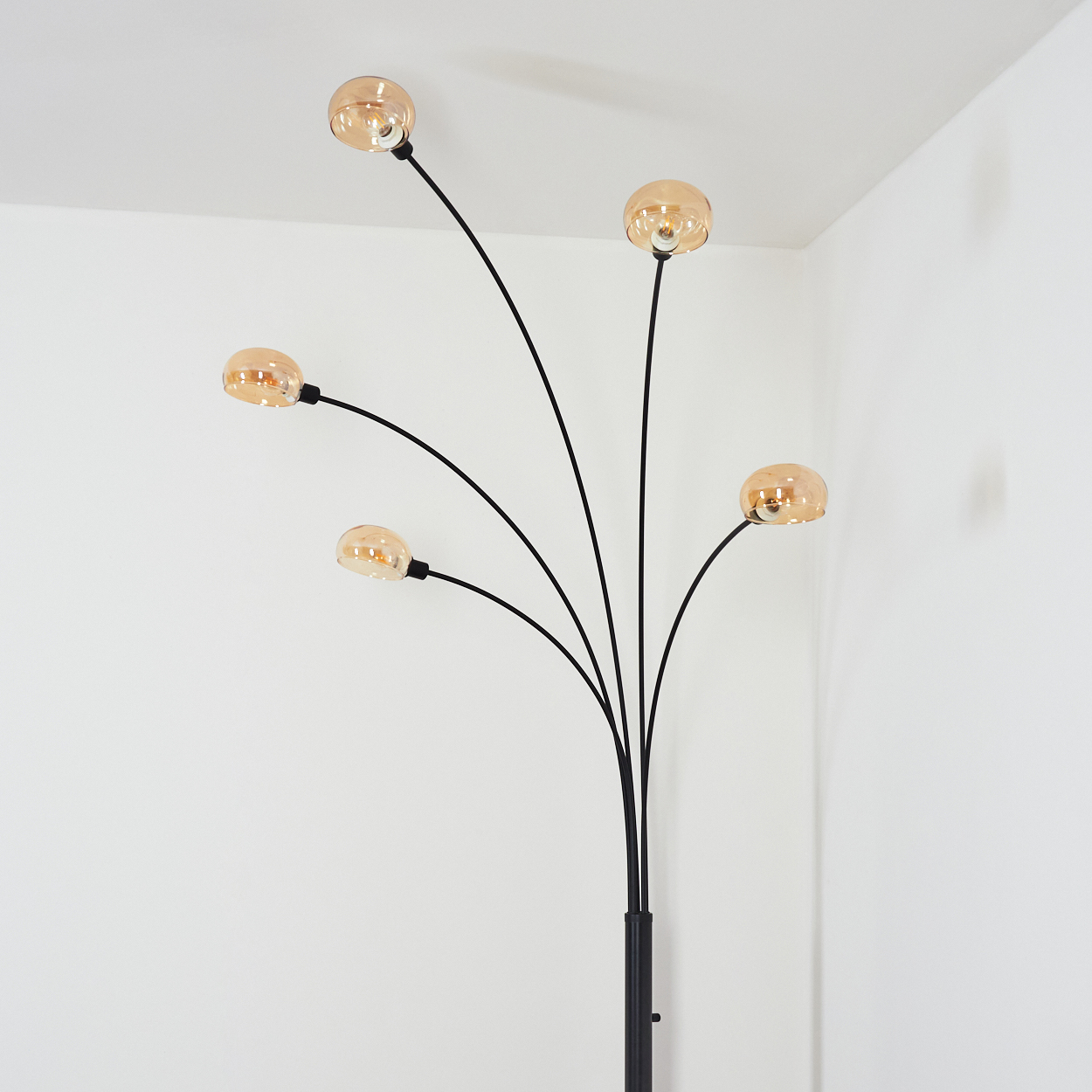 Eine moderne schwarze Stehlampe mit fünf bernsteinfarbenen Glaskugeln. Die Lampe steht in einer weißen Ecke, mit weißen Wänden und Decke.