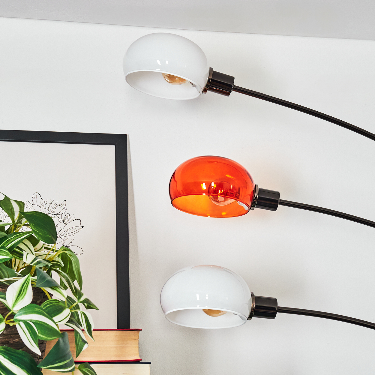 Moderne Lampe mit drei Lichtern, zwei weißen und einem orangen, vor einer weißen Wand. Eine Pflanze und Bücher im Vordergrund.
