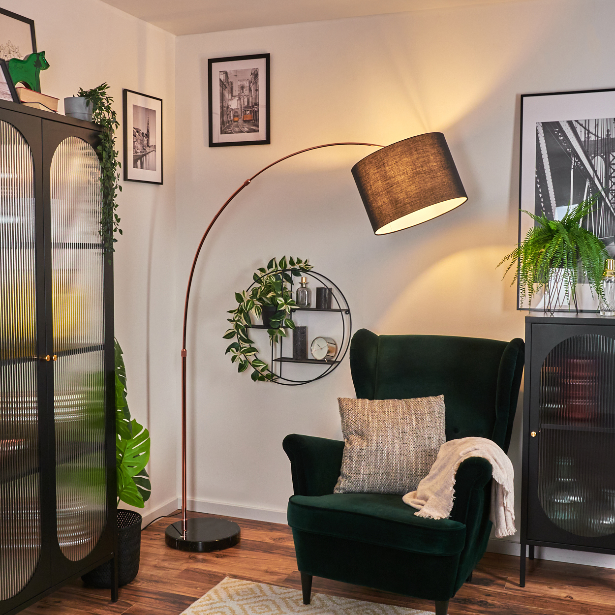 Wohnzimmerszene mit dunkelgrünem Sessel, Stehlampe und schwarzem Schrank. Pflanzen und Bilder schmücken die Wände und schaffen eine gemütliche Atmosphäre.