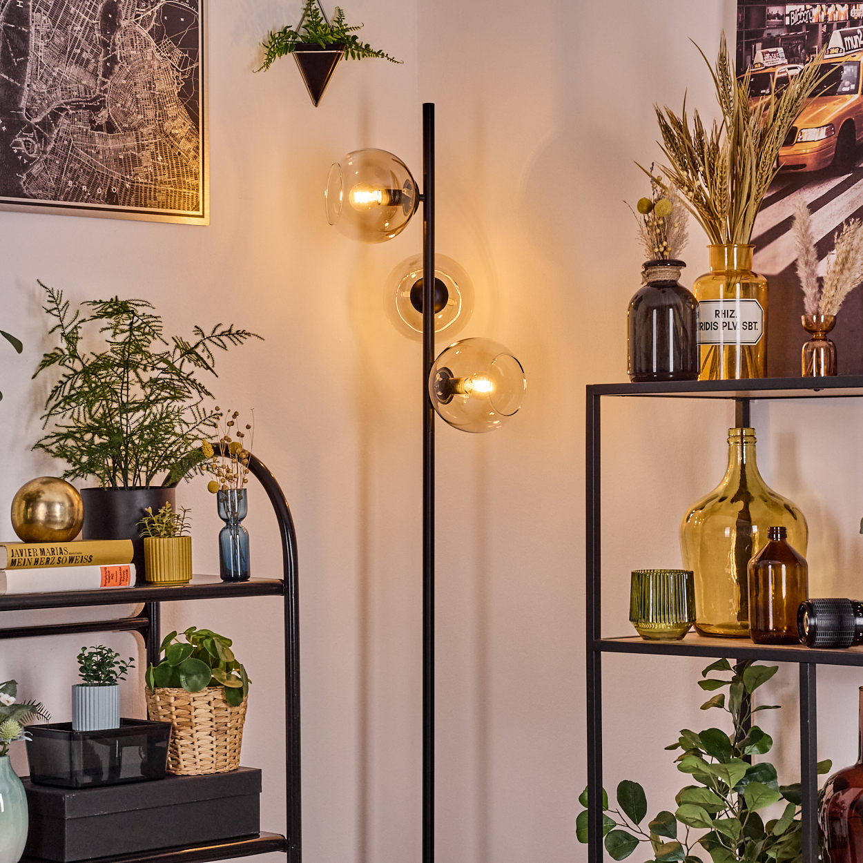Eine Zimmerecke mit einer Stehlampe mit drei Glühbirnen, Regalen mit Pflanzen und Dekorationsartikeln und einem Karten-Druck.