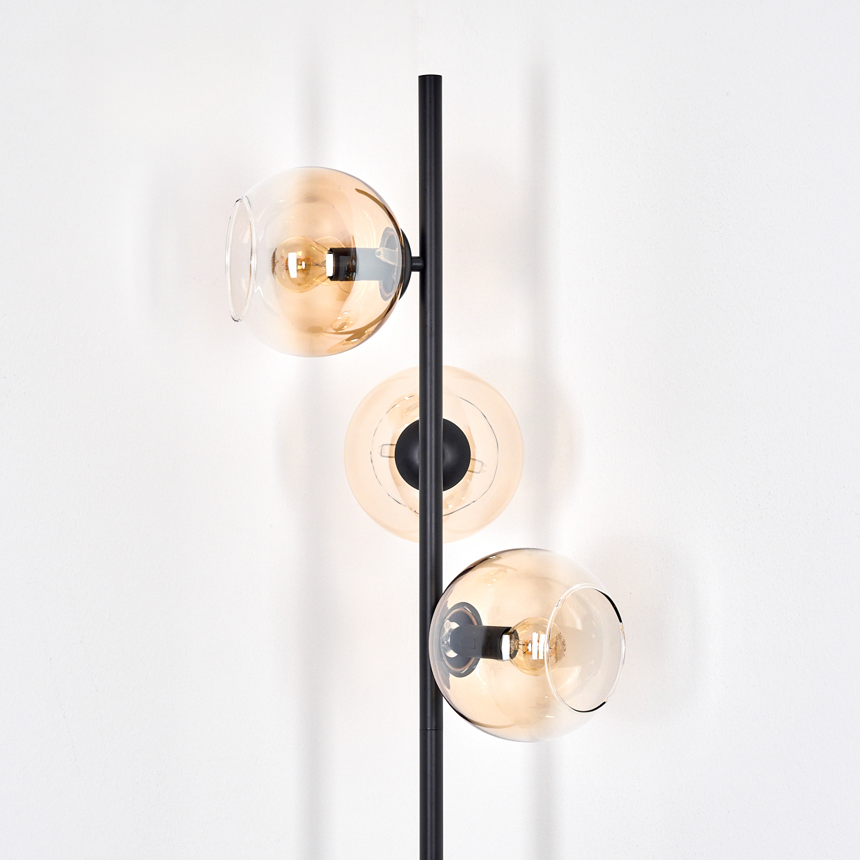 Eine moderne schwarze Stehlampe mit drei kugelförmigen, bernsteinfarbenen Glasschirmen, jeweils mit einer Glühbirne, vor einer weißen Wand.