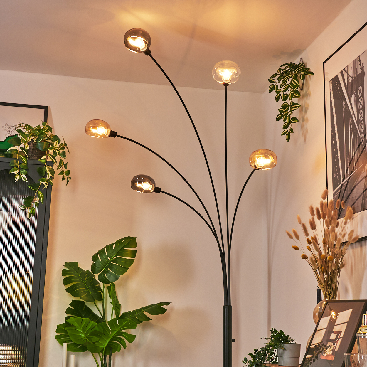 Eine moderne Stehlampe mit fünf Kugellampen. Pflanzen und ein gerahmtes Bild befinden sich im Hintergrund. Insgesamt warme, einladende Beleuchtung.
