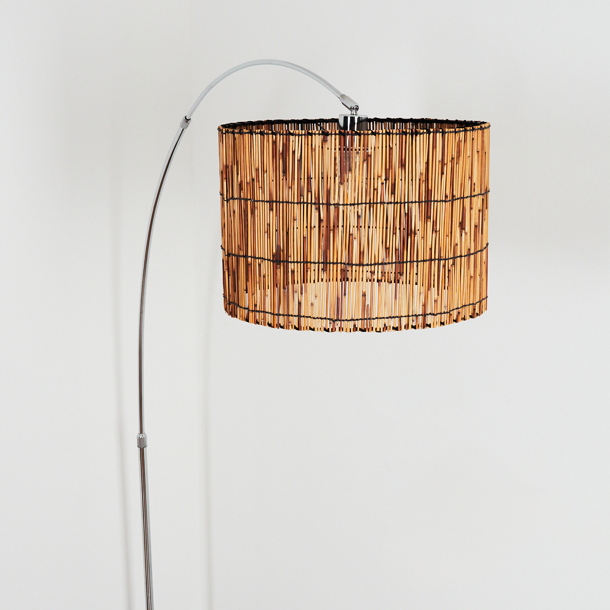Eine Stehlampe mit Bambus-Schirm. Die Lampe hat einen silbernen, gebogenen Arm und einen Sockel. Der Hintergrund ist eine weiße Wand.