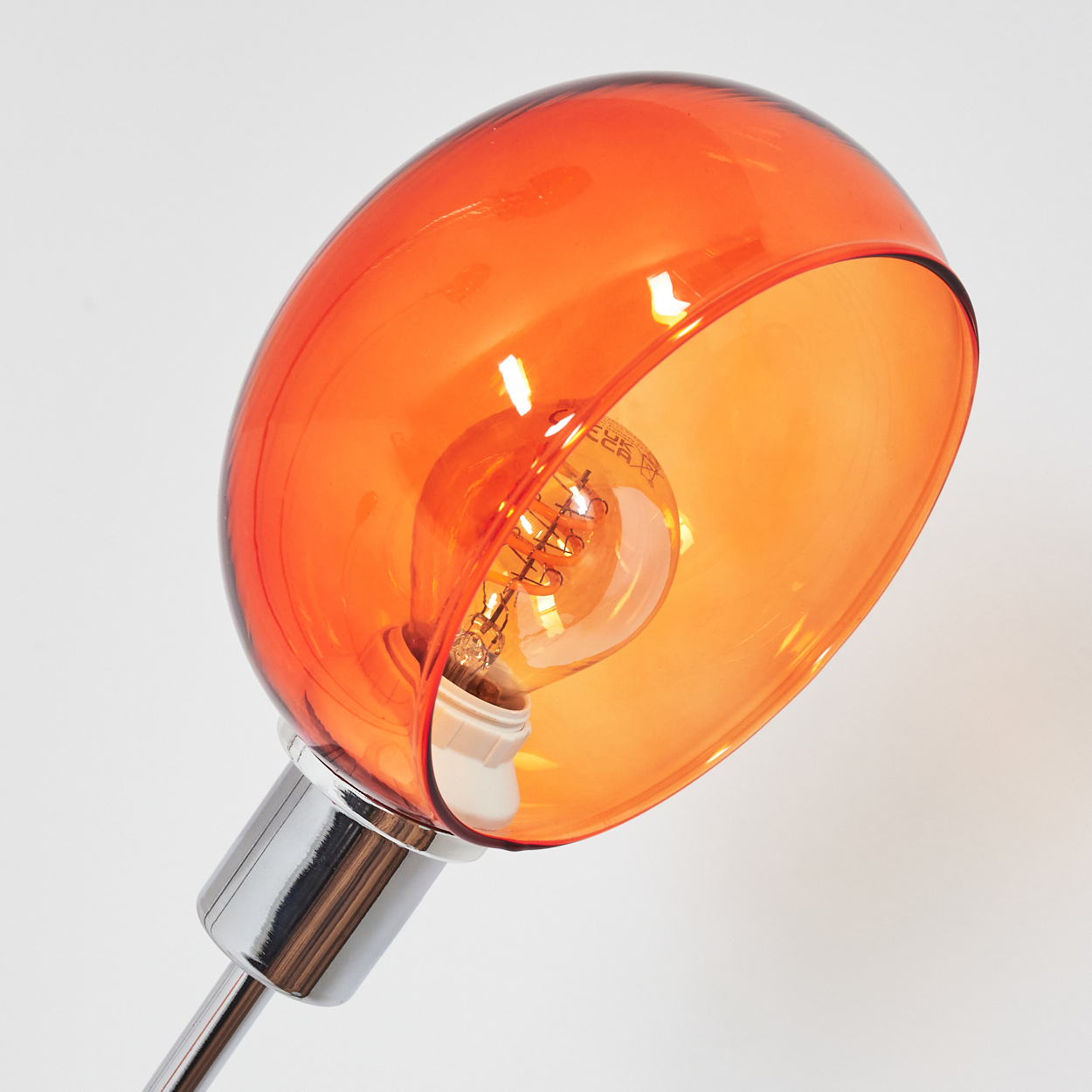 Ein orangefarbener Glasschirm mit einer Glühbirne im Inneren. Der Sockel ist verchromt, vor weißem Hintergrund.