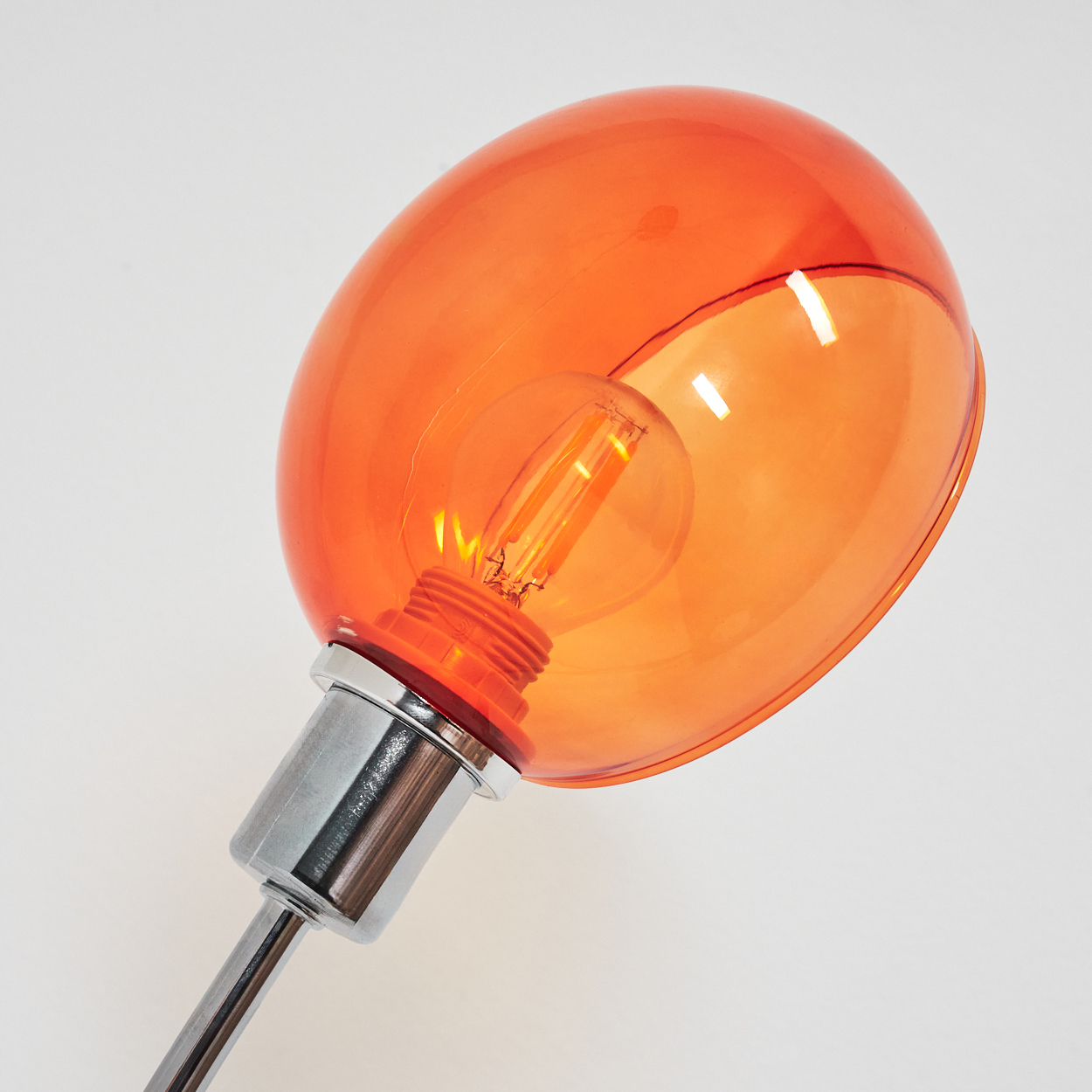 Eine orangefarbene Lampe mit einer klaren Glühbirne im Inneren. Die Lampe steht auf einem Chromschaft vor weißem Hintergrund.