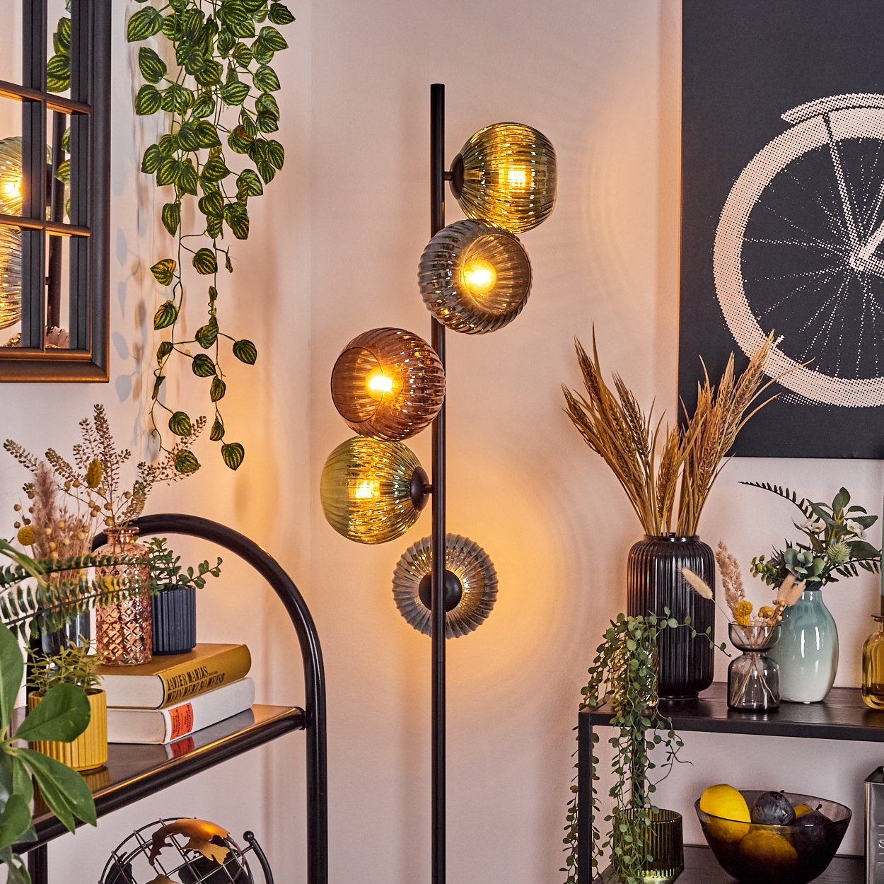 Ein modernes Wohnzimmer mit einer Stehlampe mit Glaskugeln, Pflanzen, Regalen und Dekorationsartikeln. Ein Fahrradrad-Kunstwerk hängt.