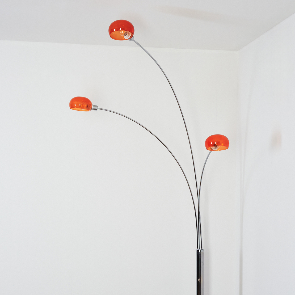 Eine moderne Stehlampe mit drei orangefarbenen Lampenschirmen und einem silbernen Rahmen vor einer weißen Wand.