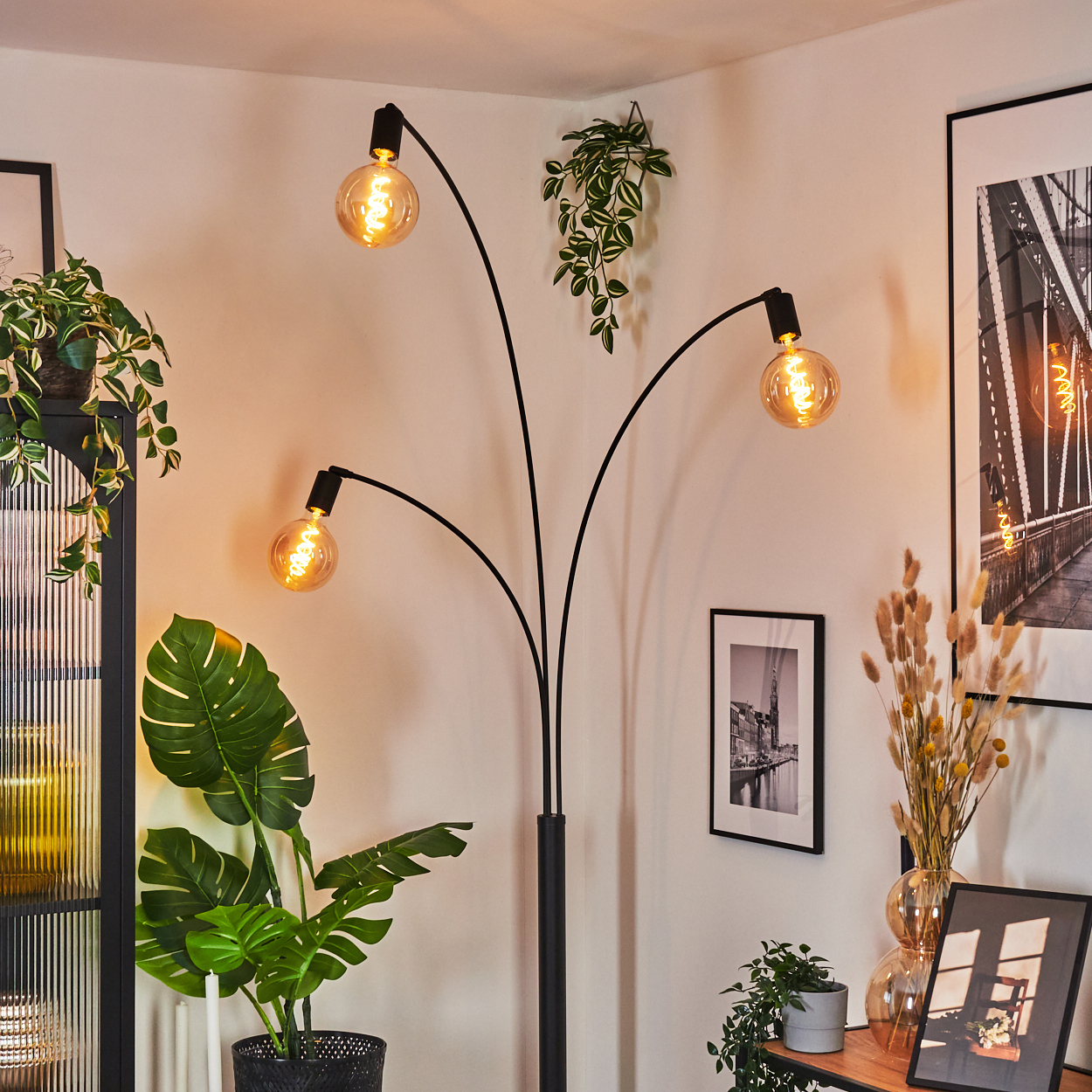 Ein modernes Wohnzimmer mit einer schwarzen Stehlampe mit drei Glühbirnen, Pflanzen und gerahmten Bildern. Der Raum ist hell.