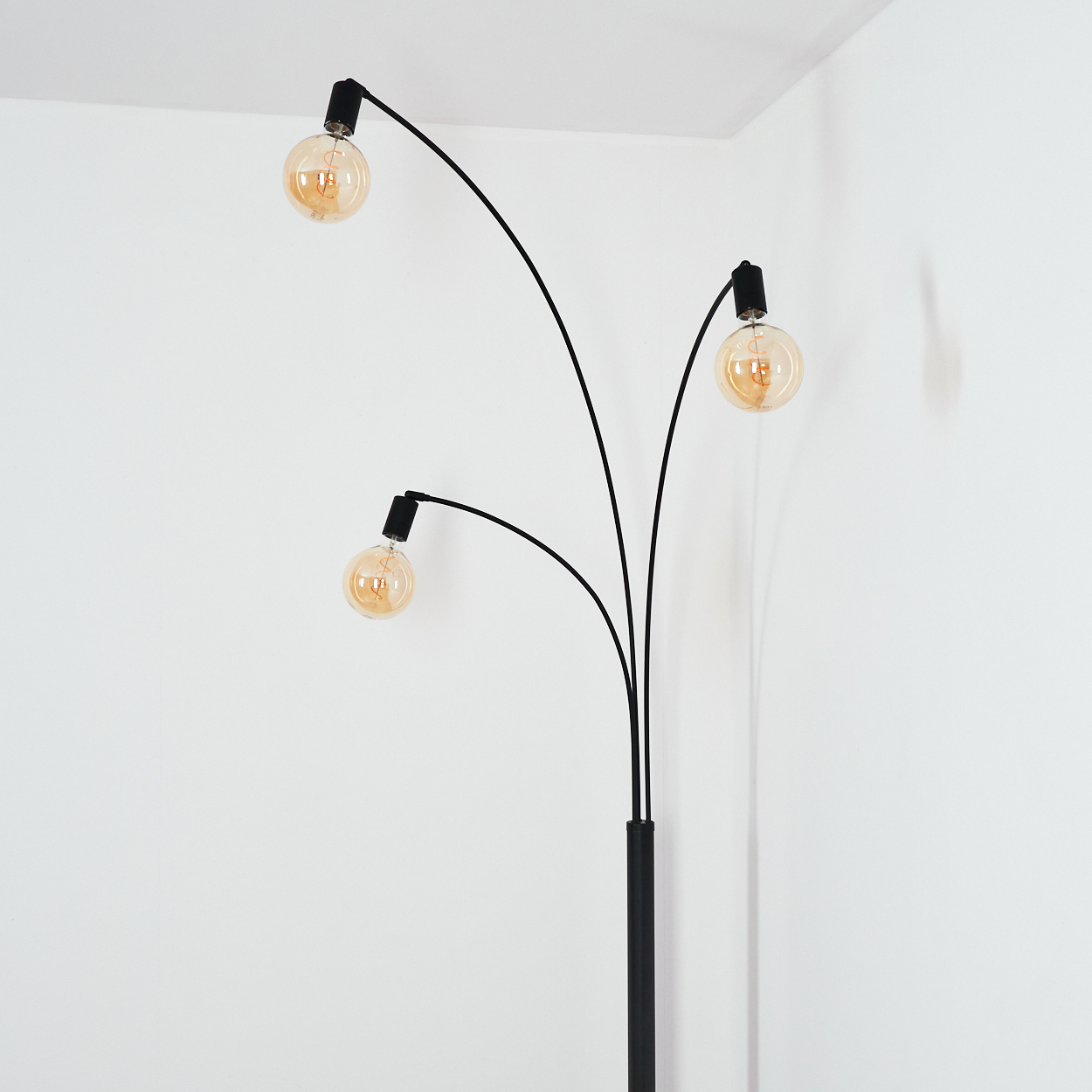 Eine schwarze Bogen-Stehlampe mit drei Glühbirnen. Die Glühbirnen sind rund und goldfarben, vor einer weißen Wand.