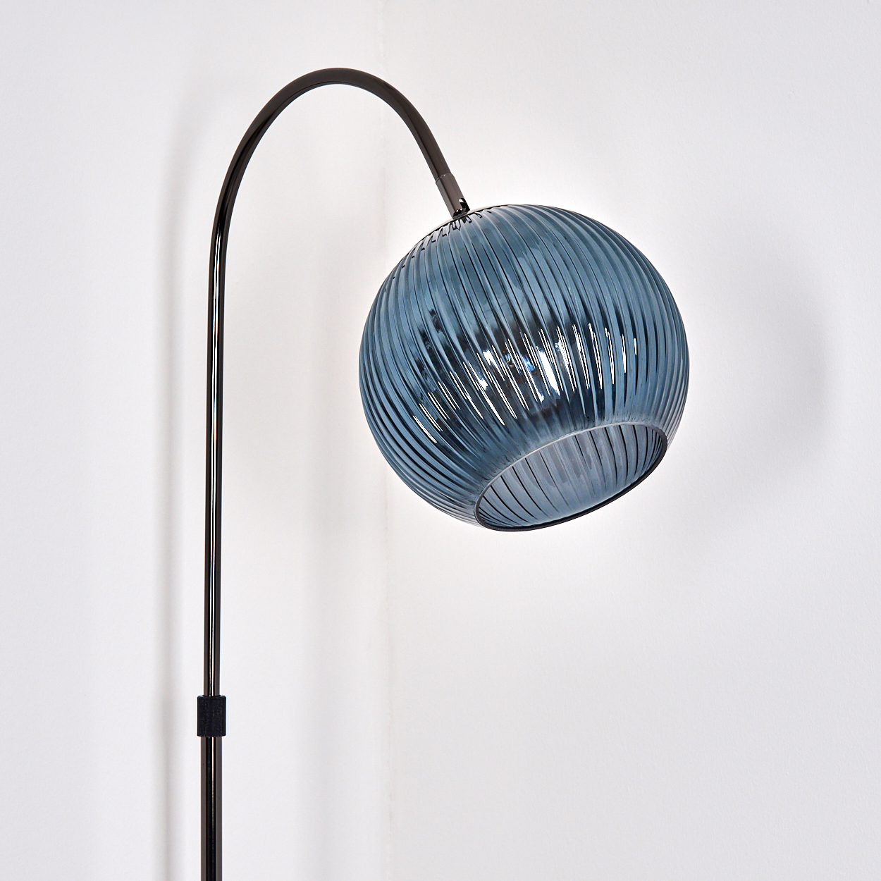 Eine moderne Stehlampe mit blauem Glaskugel-Schirm und schwarzem Metallrahmen vor einer weißen Wand.