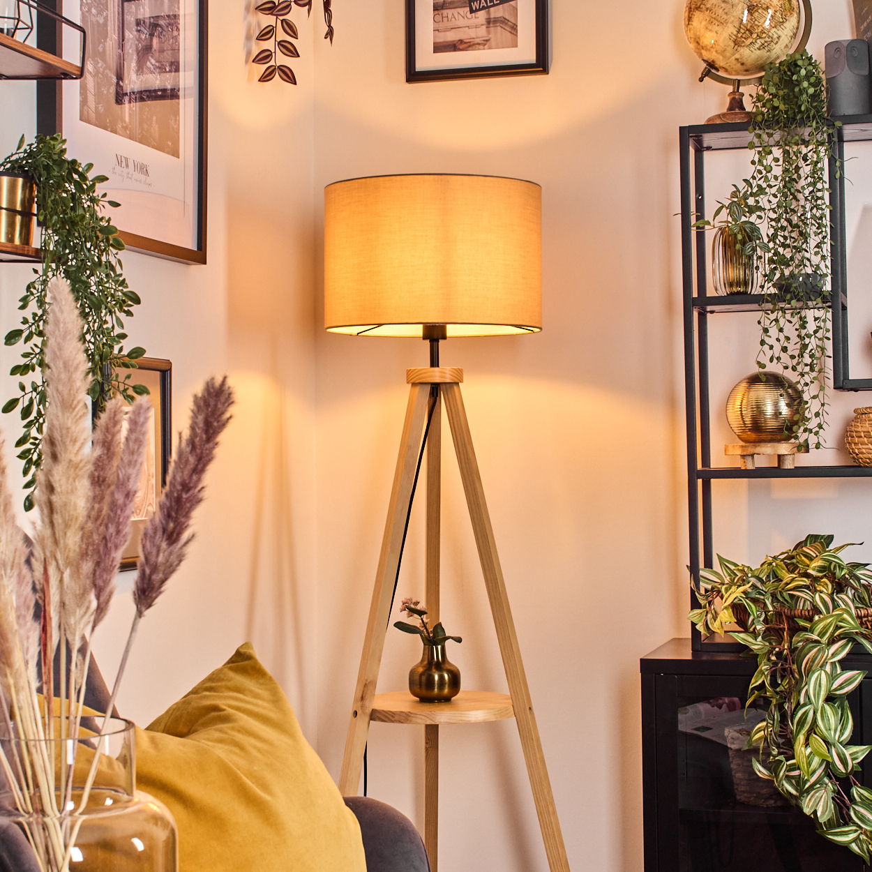 Eine beleuchtete hölzerne Stativlampe steht in einer Ecke mit Pflanzen, Bildern und Regalen. Eine goldene Vase hält eine kleine Blume.