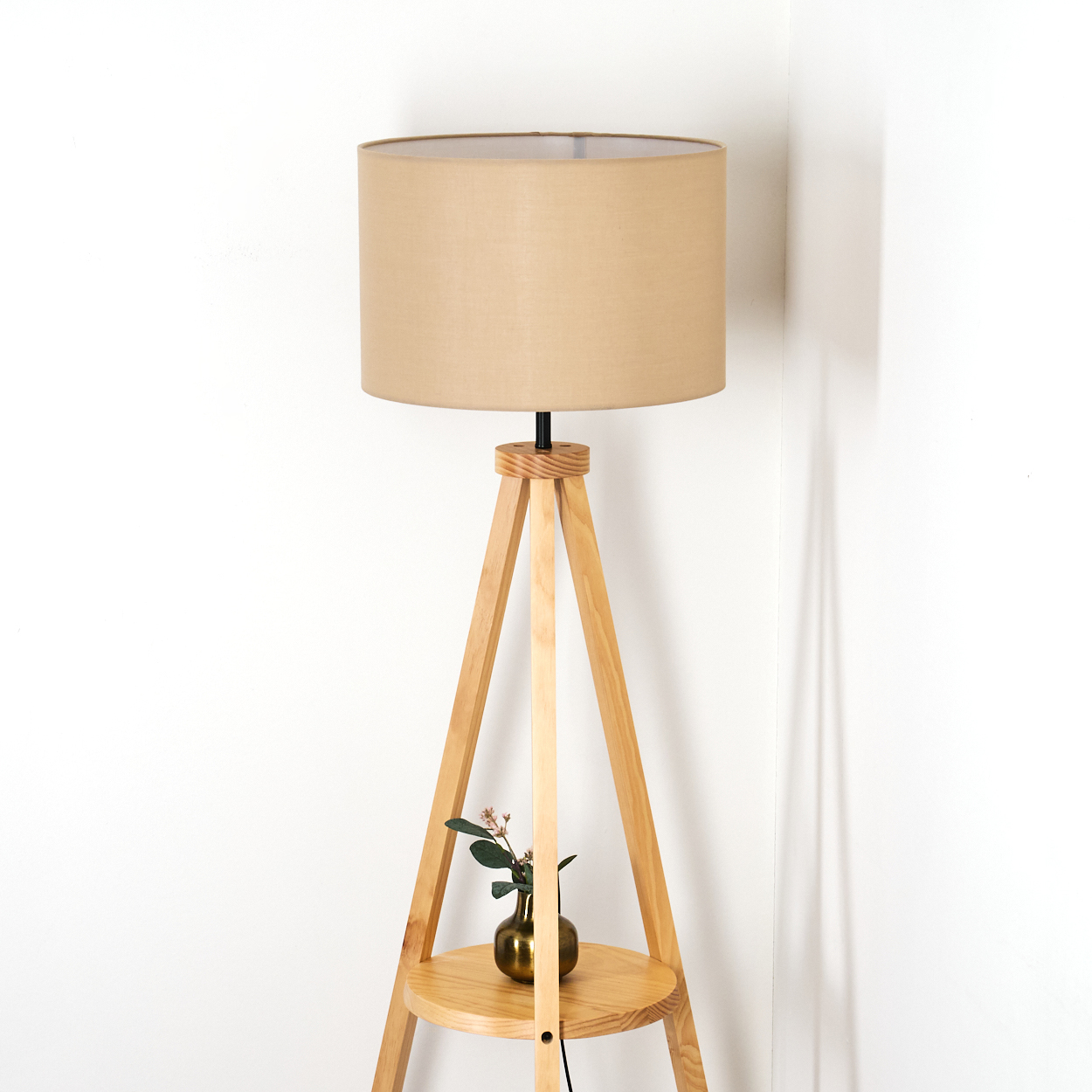 Ein beigefarbener Lampenschirm steht auf einer hölzernen Stativlampe. Eine kleine goldene Vase mit Grün steht auf einem Regal. Weißer Wandhintergrund.