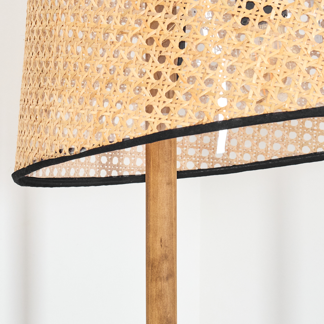 Nahaufnahme eines Lampenschirms aus Rattan mit schwarzem Rand und Holzstange. Der Hintergrund ist eine weiße Wand.