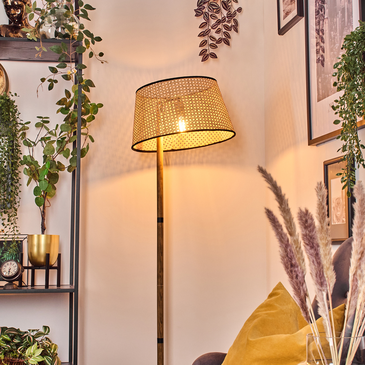 Eine beleuchtete Stehlampe mit gewebtem Schirm steht in einer Ecke. Pflanzen und Bilder schmücken die Wand. Im Vordergrund liegt ein gelbes Kissen.
