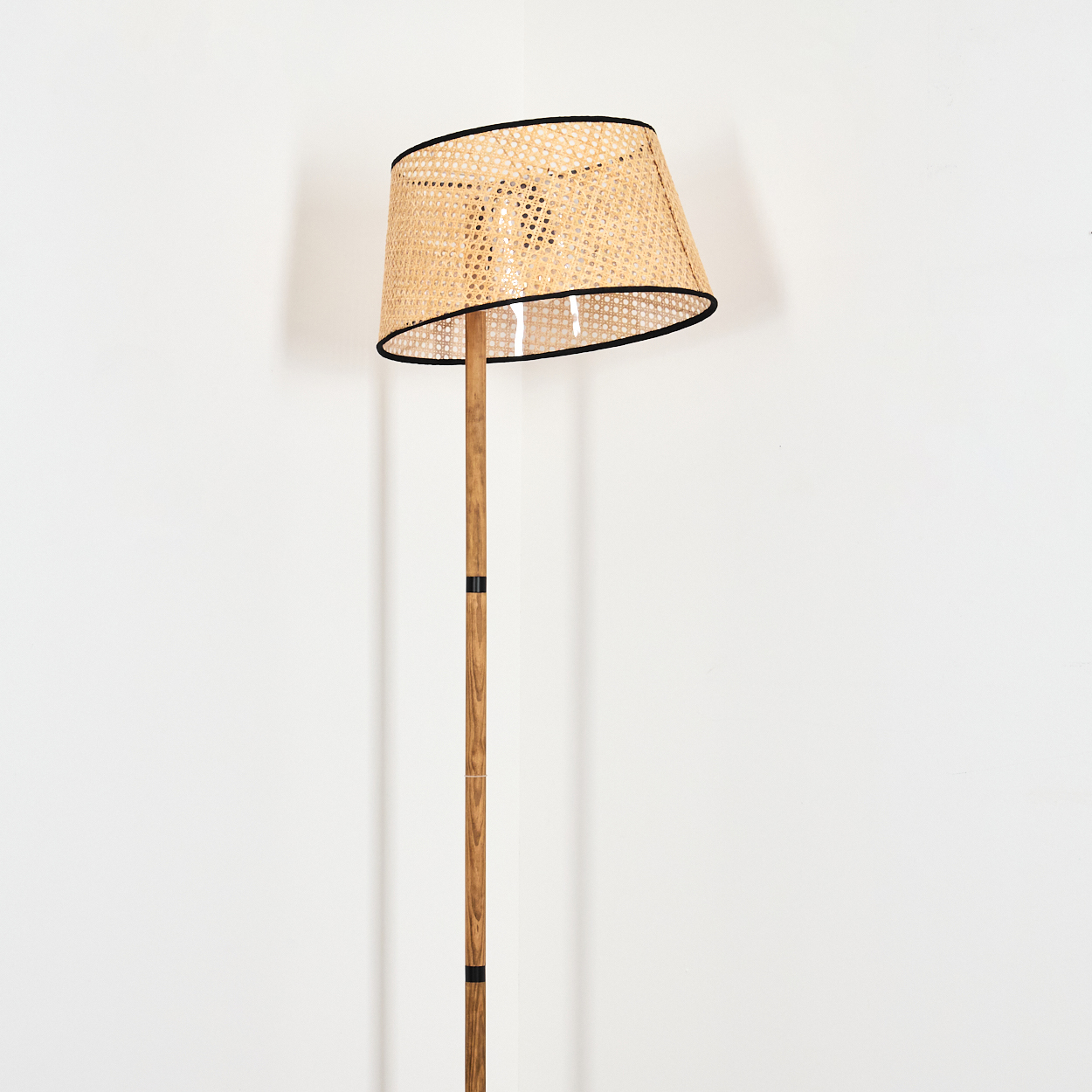 Eine Stehlampe mit gewebtem Lampenschirm und Holzstange steht vor einer weißen Wand. Der Lampenschirm hat eine schwarze Einfassung.