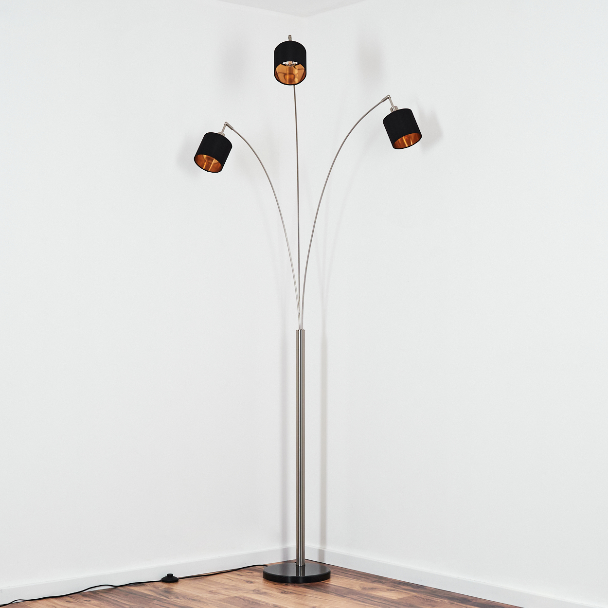 Eine moderne Stehlampe mit drei schwarzen Schirmen und einem silbernen Ständer steht vor einer weißen Wand und Holzboden.