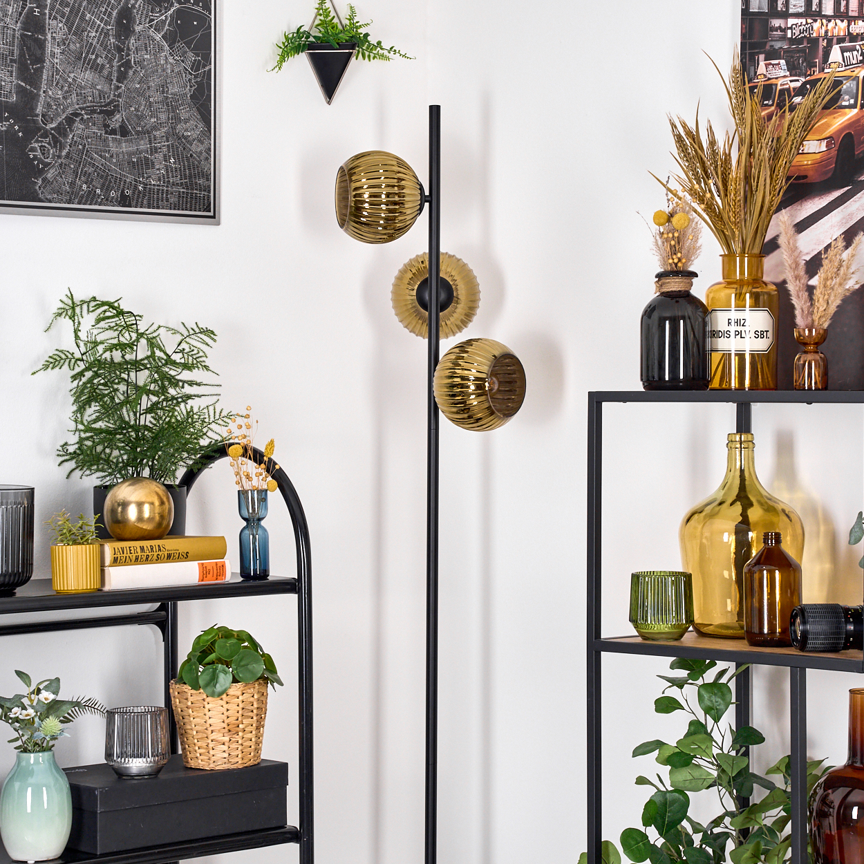 Eine Zimmerecke mit Regalen, Pflanzen und einer Stehlampe mit drei goldenen Glaskugeln. Eine Karte und ein Taxiposter an der Wand.