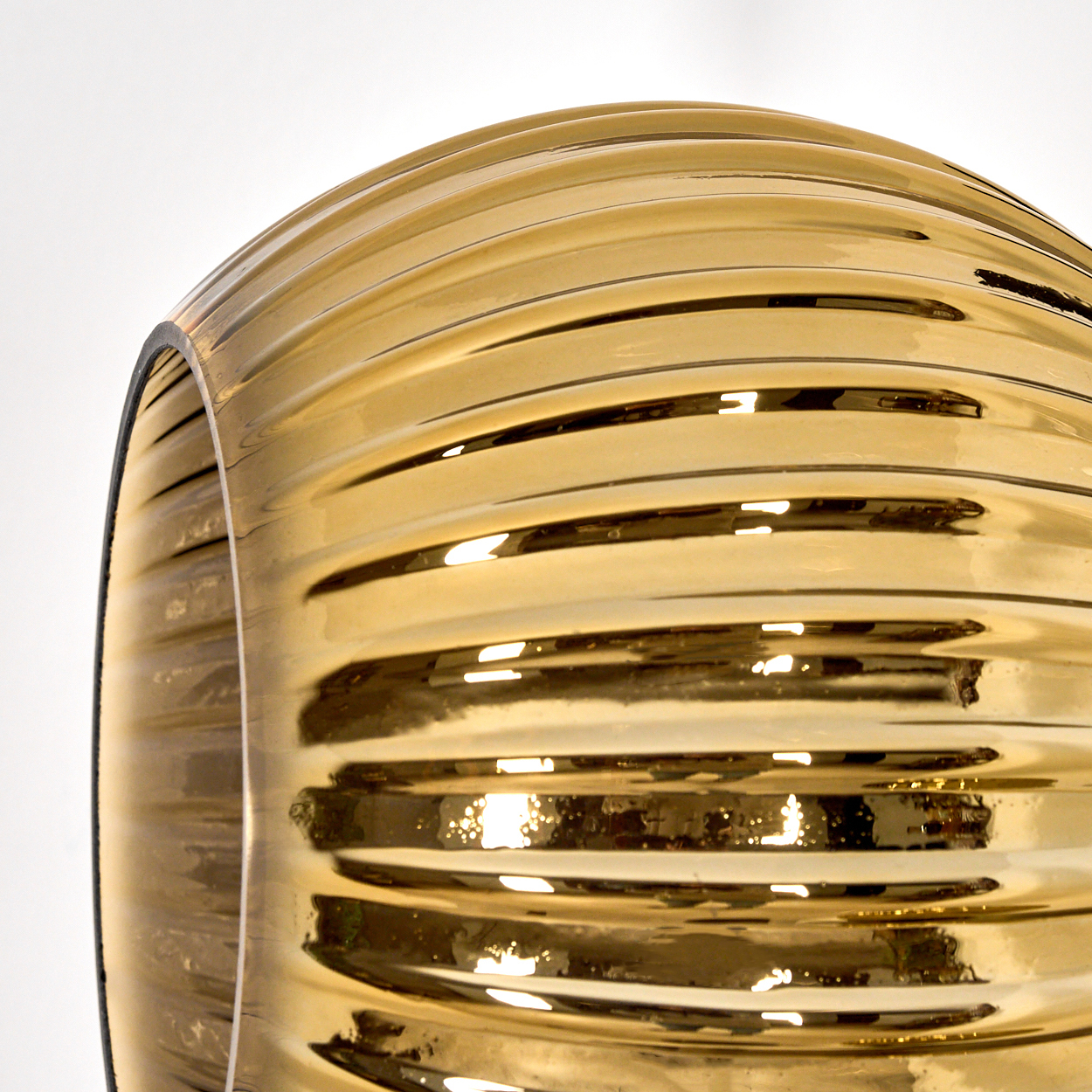 Nahaufnahme eines goldenen Glasobjekts, möglicherweise ein Lampenschirm, mit horizontalen Rippen. Der Hintergrund ist weiß.