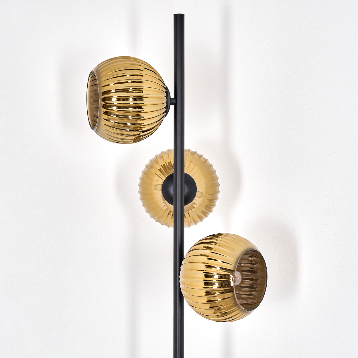 Eine moderne Lampe mit schwarzem Pfosten und drei goldenen, gerippten Glasschirmen. Der Hintergrund ist eine weiße Wand.