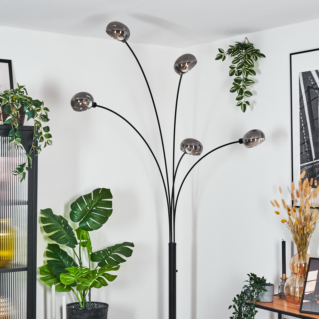 Eine moderne Stehlampe mit fünf Lichtern. Im Hintergrund befinden sich Pflanzen und Dekorationen. Die Wand ist weiß.