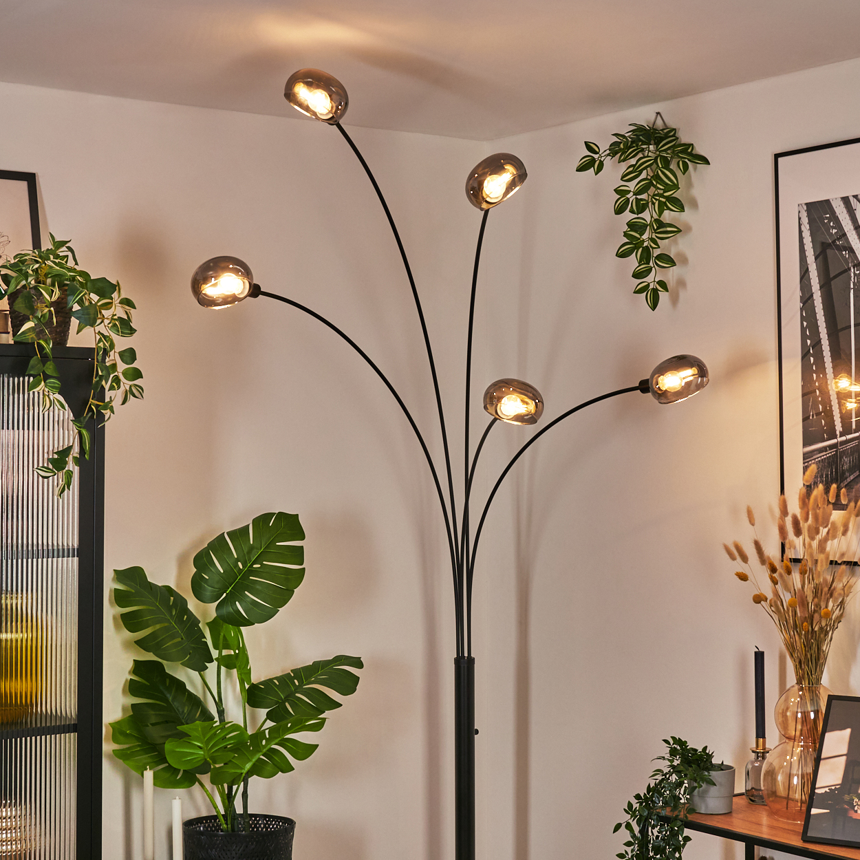 Ein modernes Wohnzimmer mit einer schwarzen Stehlampe mit fünf Lichtern. Pflanzen und Bilder schmücken den Hintergrund.