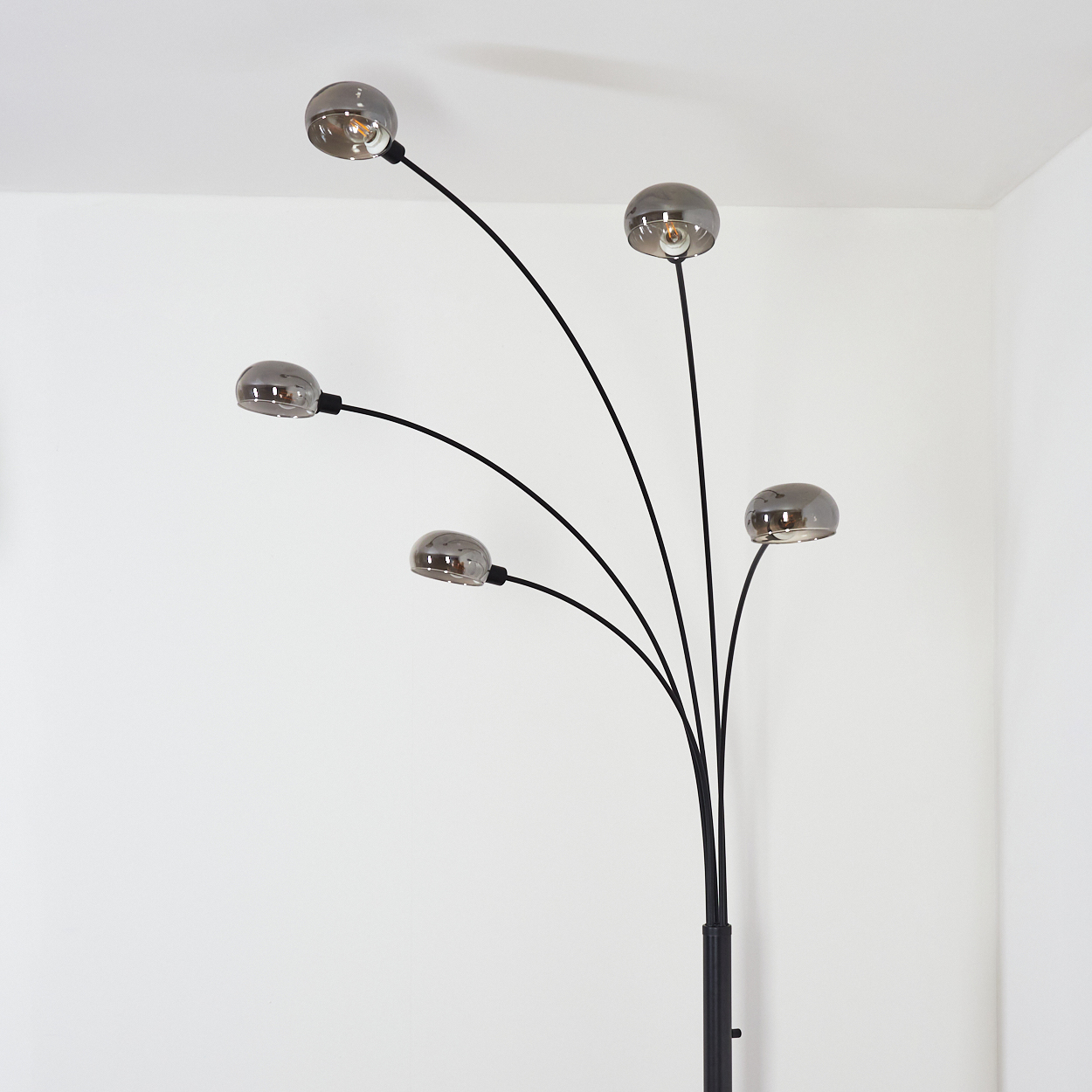 Eine moderne Stehlampe mit fünf kugelförmigen Lichtern. Die Lampe hat einen schwarzen Stiel und Arme mit Milchglasschirmen vor weißer Wand.