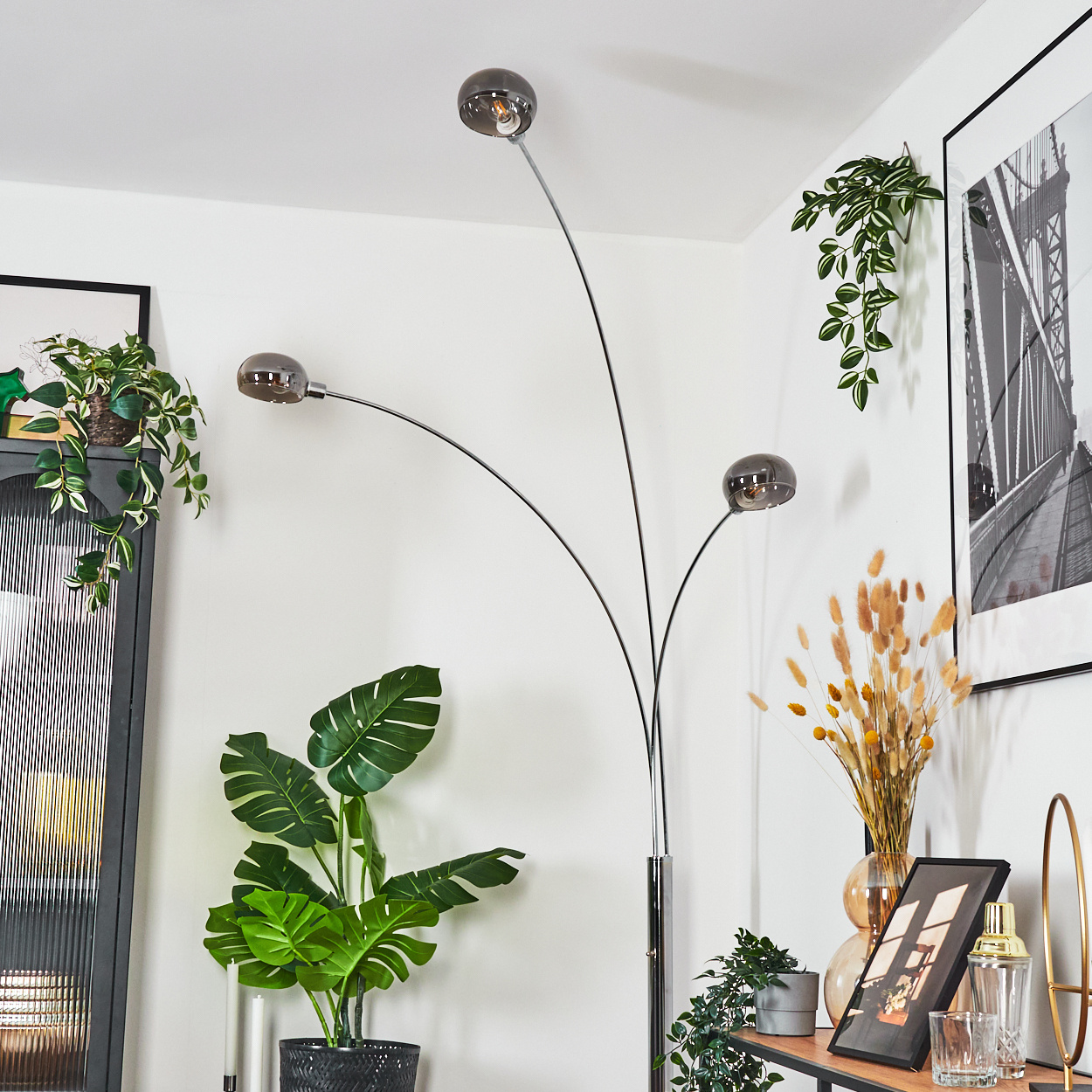 Ein modernes Wohnzimmer mit Stehlampe, Pflanzen und gerahmter Kunst. Die Lampe hat drei Kugelleuchten. Pflanzen stehen in schwarzen Töpfen.