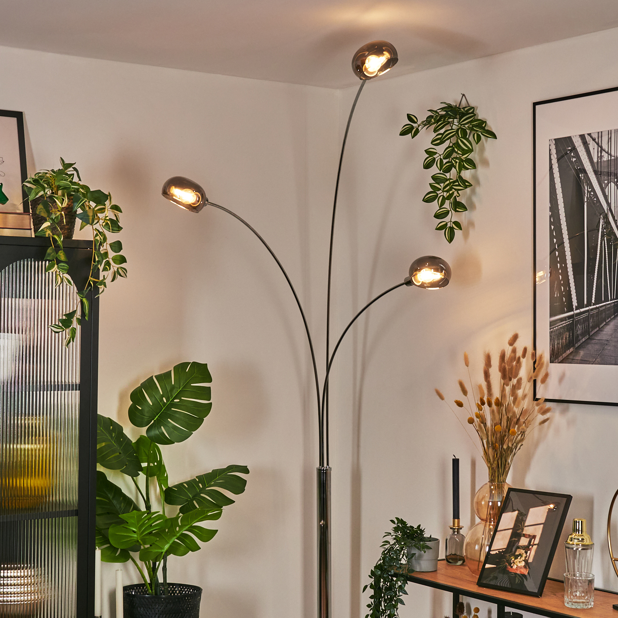Ein modernes Wohnzimmer mit Stehlampe, Pflanzen und einem Bild an der Wand. Die Lampe hat drei Leuchten.