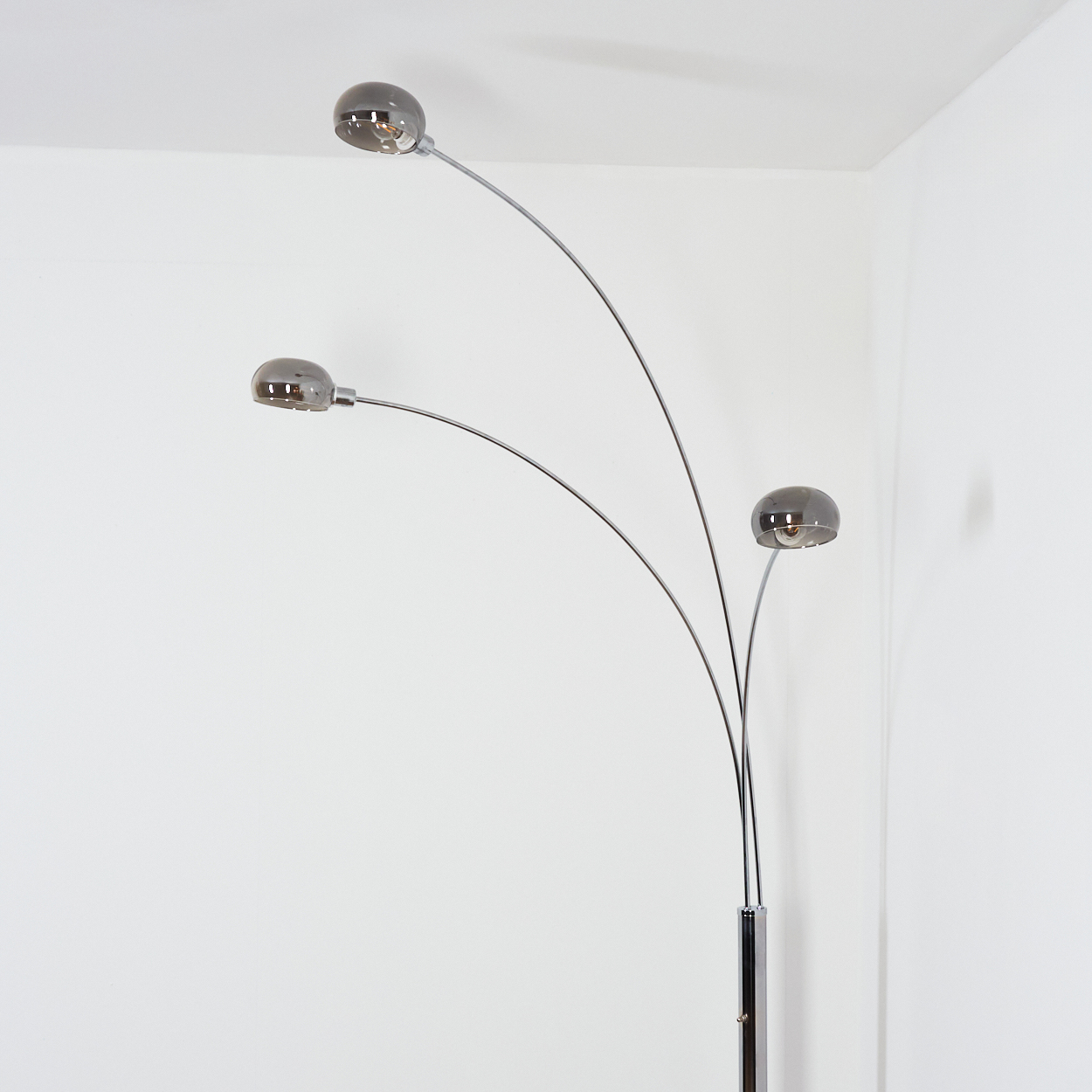 Eine moderne Stehlampe mit drei gebogenen Armen, die jeweils eine kugelförmige Leuchte aus Rauchglas halten. Die Lampe steht vor einer weißen Wand.