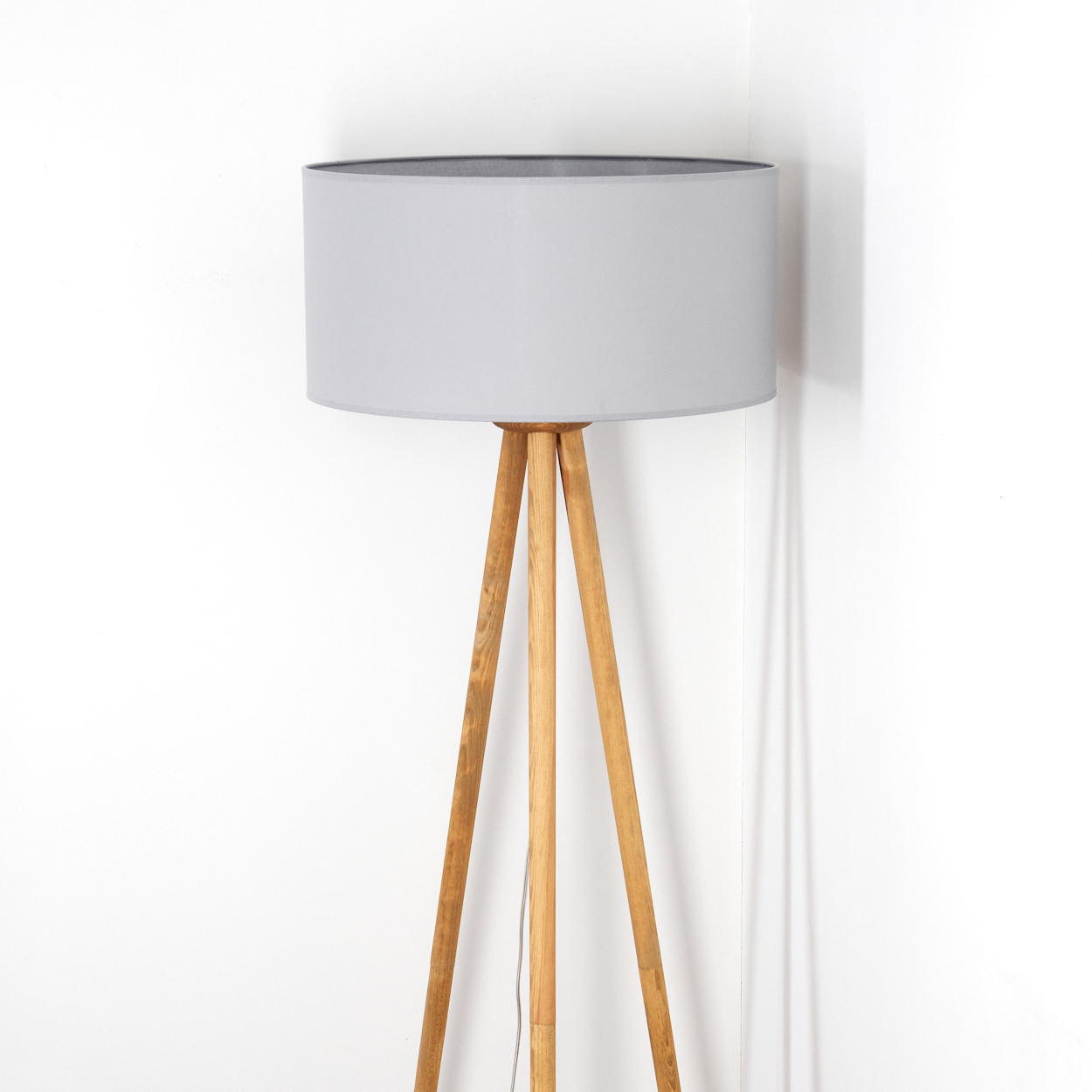 Eine Stehlampe steht in einer Ecke. Sie hat einen grauen Lampenschirm und Holzstative. Der Hintergrund ist eine weiße Wand.
