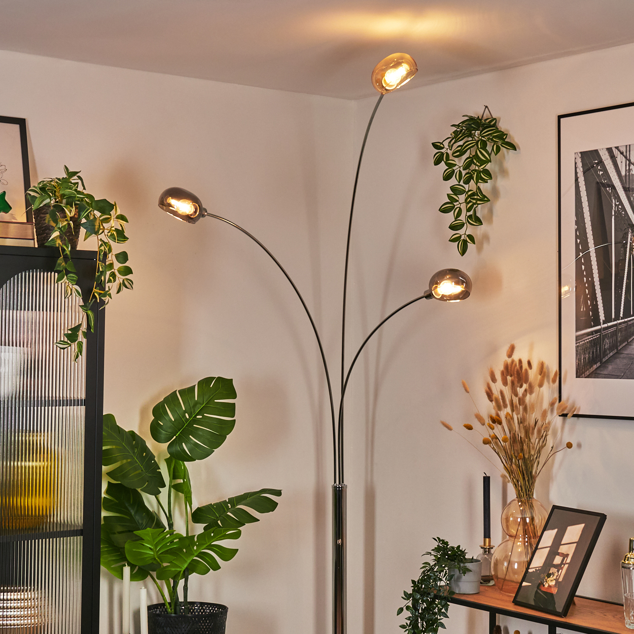 Eine moderne Stehlampe mit vier Lichtern. Pflanzen und Bilder schmücken den Raum. Warmes Licht erhellt die Szene.