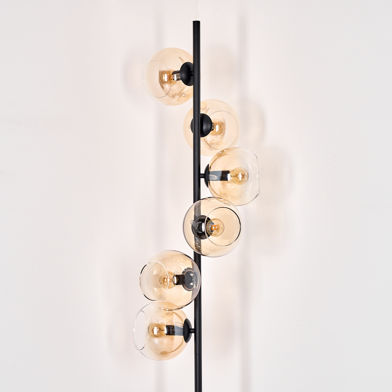 Eine moderne Stehlampe mit sechs bernsteinfarbenen Glaskugeln. Der schwarze Metallrahmen hält die Kugeln an einer weißen Wand.