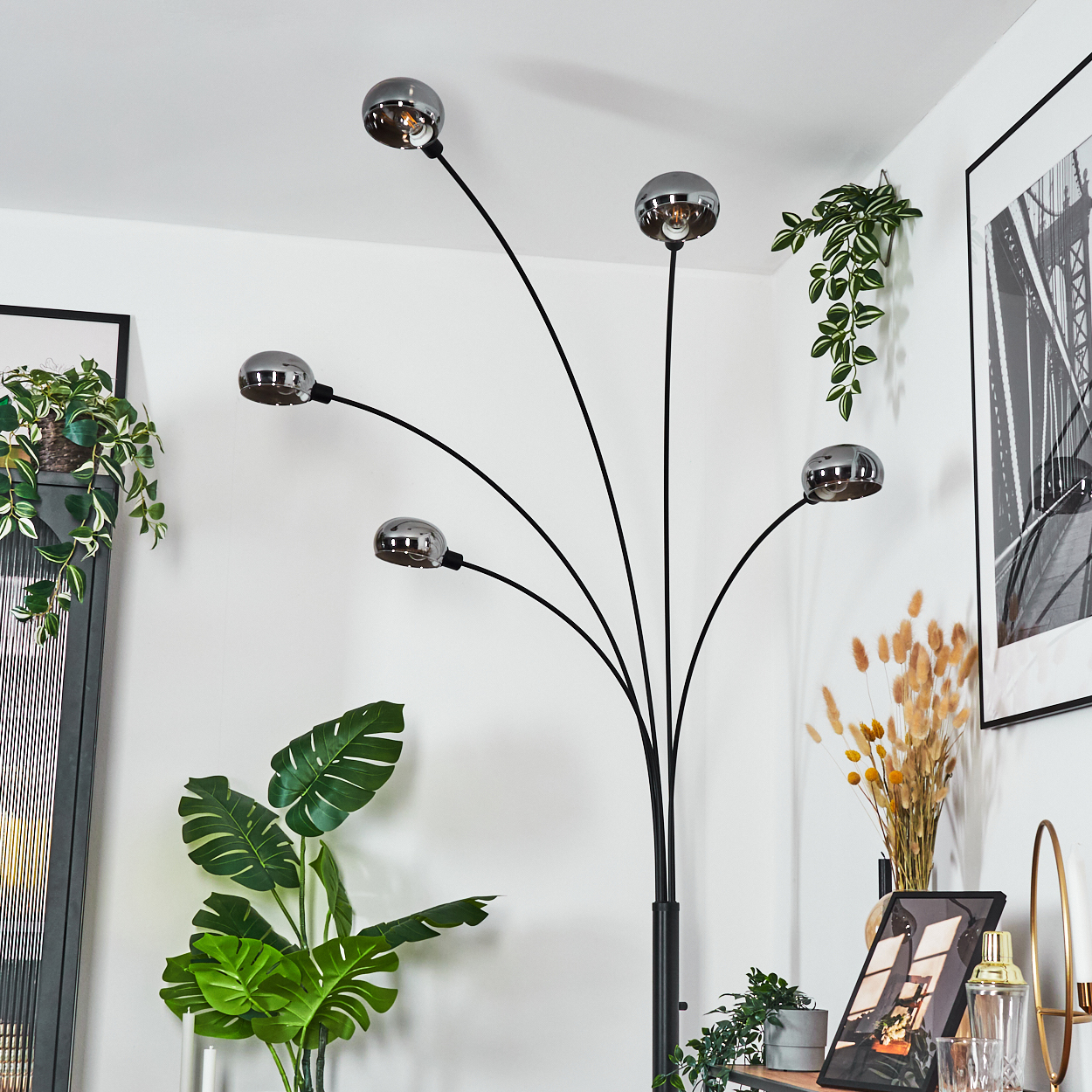 Ein modernes Wohnzimmer mit einer schwarzen Bogenlampe, Pflanzen und gerahmten Kunstwerken. Die Lampe hat fünf Kugelleuchten. Pflanzen sind grün.